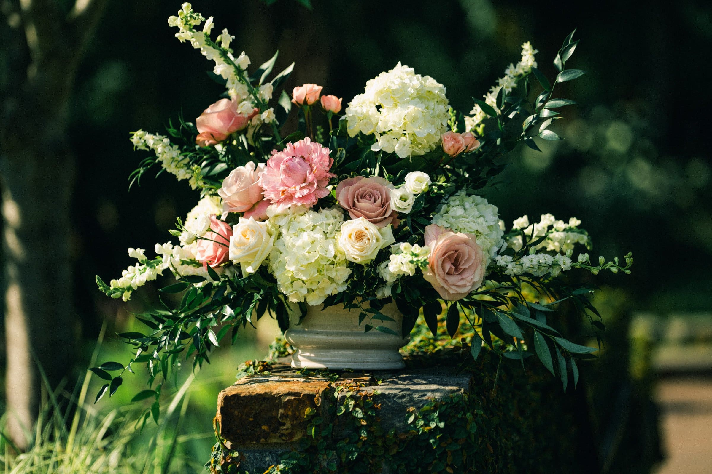 duke gardens wedding photography - ann & tim A floral arrangement in a white vase featuring pink roses, white hydrangeas, and lush green foliage graces a stone ledge. Perfect for Duke Gardens wedding photography, it captures the essence of romance against a soft-focus garden backdrop.