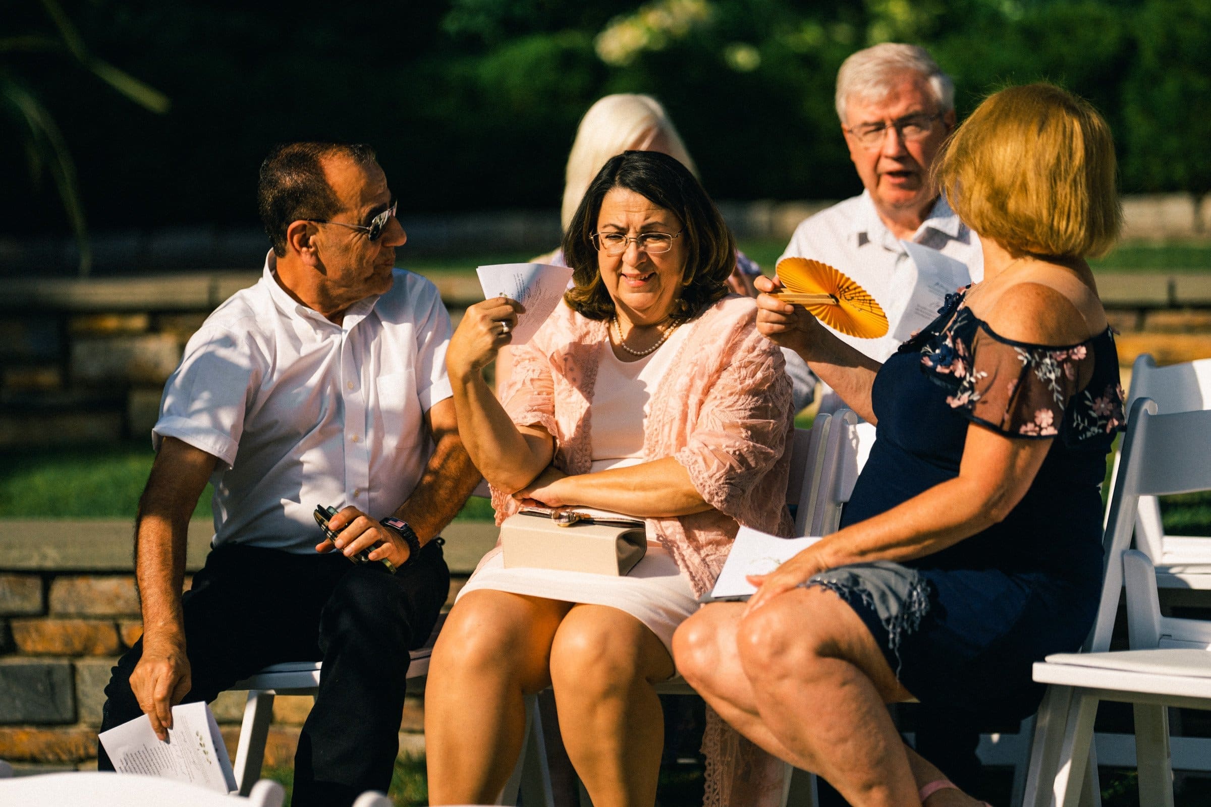 duke gardens wedding photography - ann & tim A group of four older adults, two men and two women, sit outdoors on white chairs amidst the serene beauty reminiscent of Duke Gardens, engaging in conversation. One woman fans herself with a paper fan. They seem relaxed under sunlight, with greenery in the background.