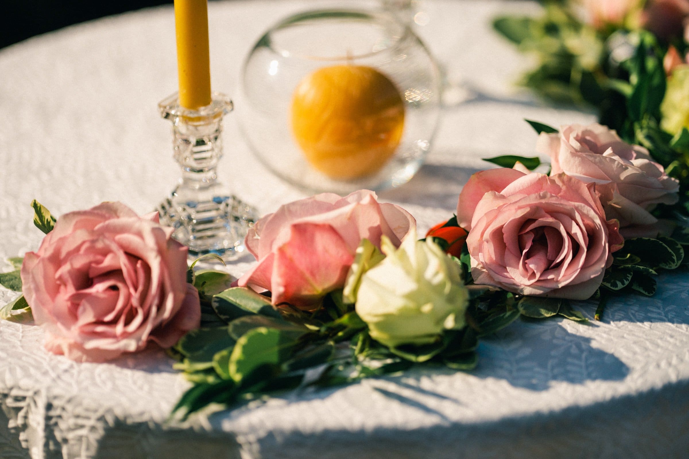 duke gardens wedding photography - ann & tim A white lace tablecloth adorned with pink and white roses, green leaves, a clear candle holder with a yellow candle, and a round glass vase with a lemon inside evokes the charm of Duke Gardens wedding photography, creating an elegant decorative arrangement.