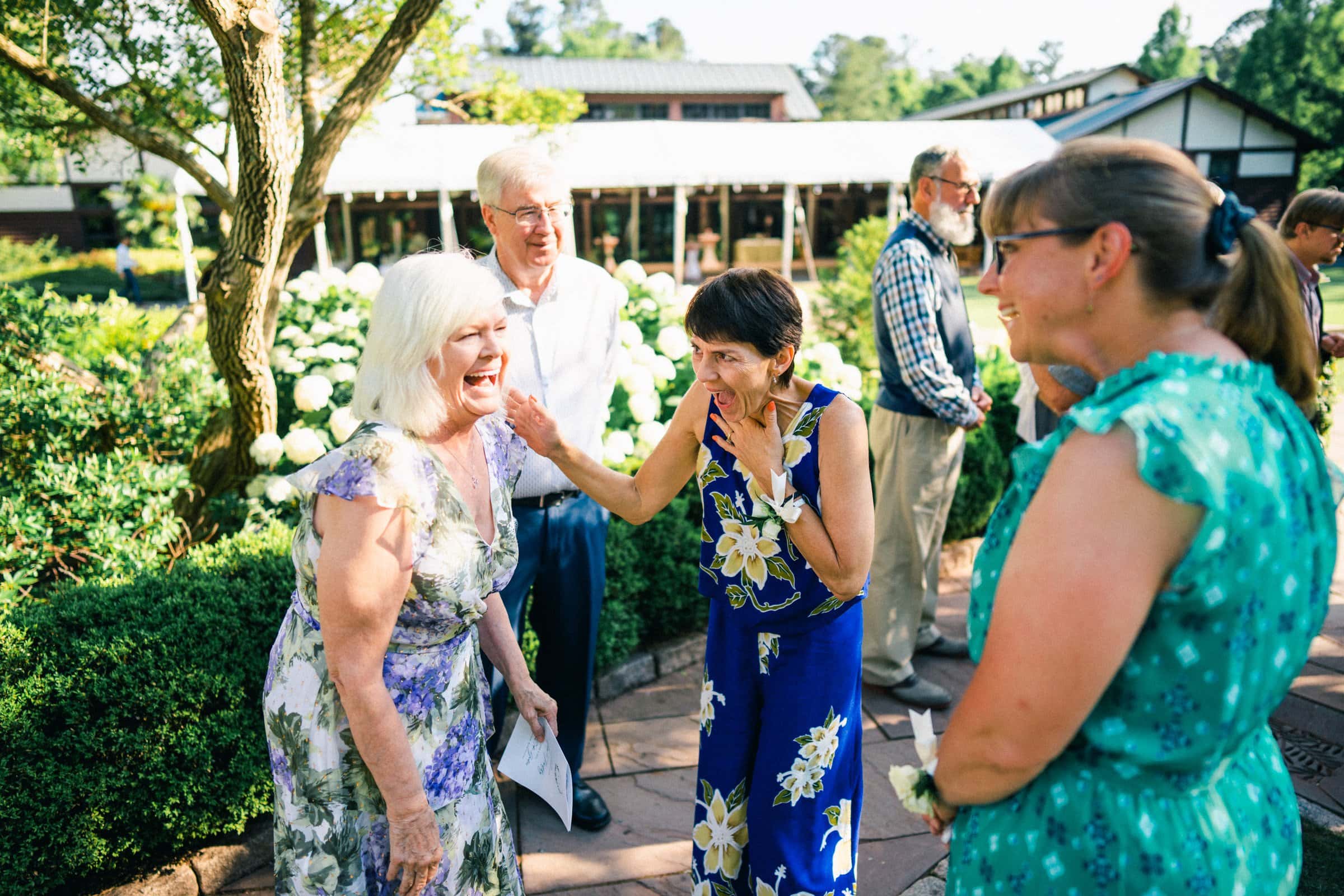 duke gardens wedding photography - ann & tim A group of people outdoors are smiling and laughing together, reminiscent of a joyful gathering at Duke Gardens. The focus is on an older woman with white hair in a floral dress sharing laughter with another woman in a blue floral outfit, as others in the background enjoy cheerful conversation, perfectly captured in wedding photography style.