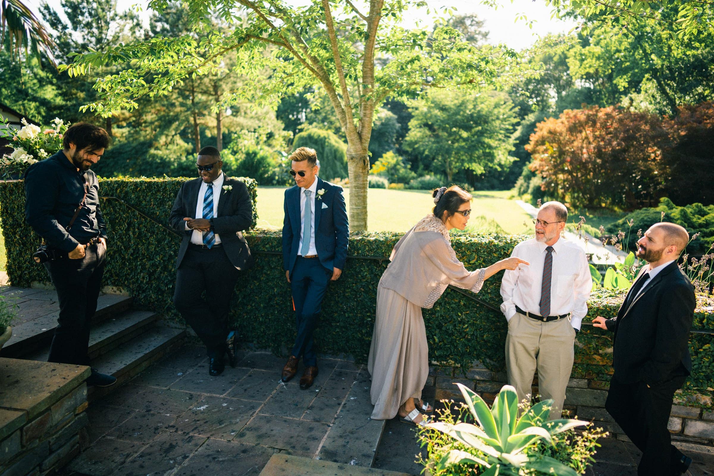 duke gardens wedding photography - ann & tim In a garden setting perfect for wedding photography, a group of six—four men and two women—are elegantly dressed in formal attire. They stand or sit casually near a stone ledge at Duke Gardens, surrounded by lush greenery and sunlight as they engage in lighthearted conversation.