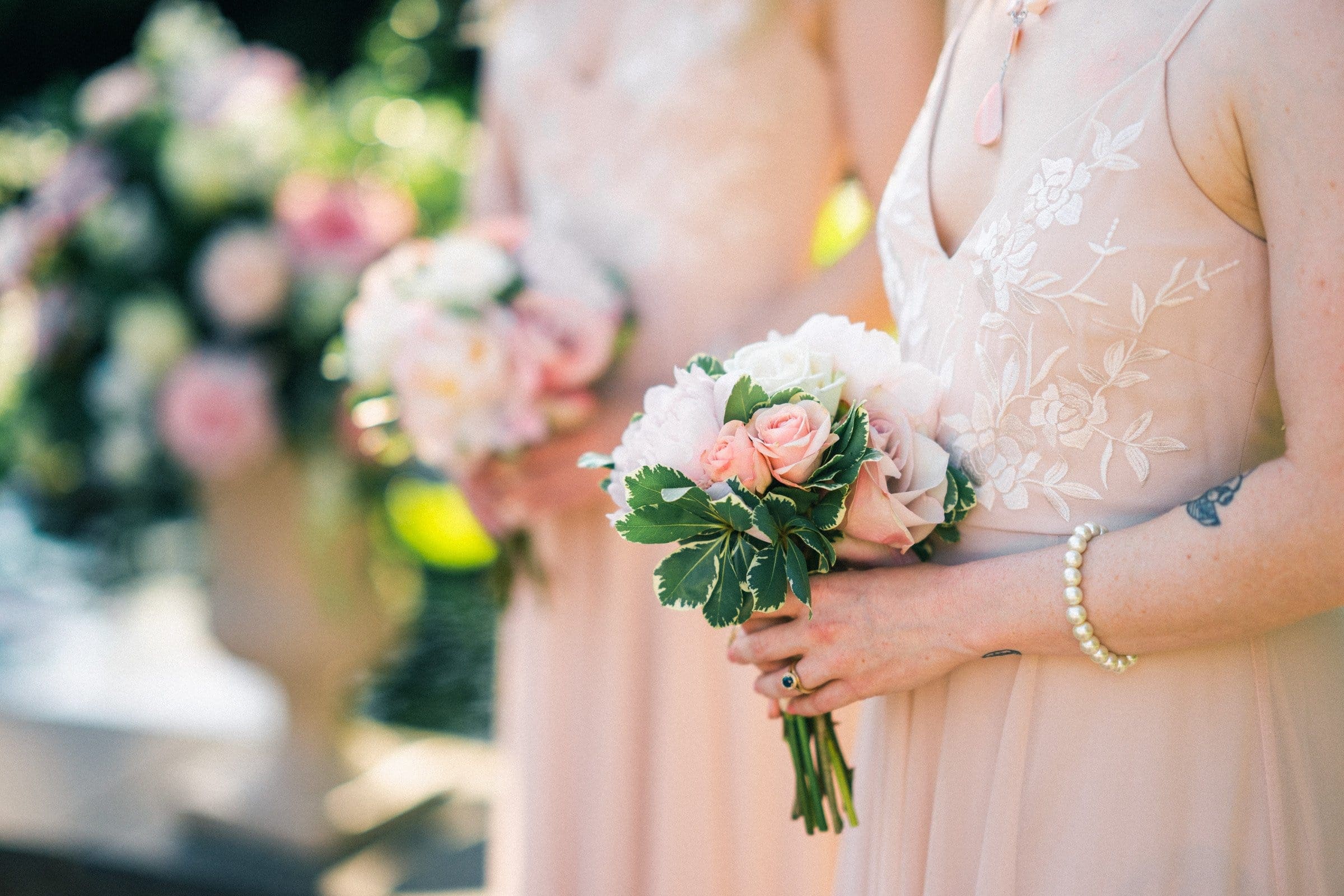 duke gardens wedding photography - ann & tim Two people in elegant, pastel-colored dresses hold bouquets of pink and white flowers. The scene, reminiscent of Duke Gardens wedding photography, is bright and features soft-focus floral arrangements in the background, suggesting a peaceful, outdoor setting.