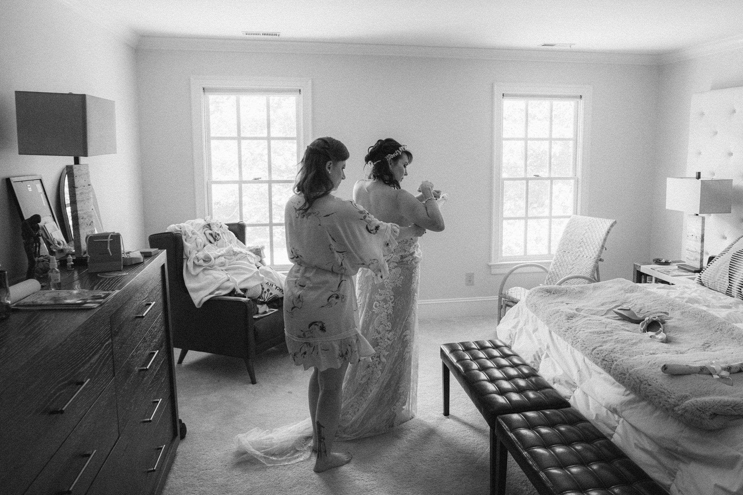 duke gardens wedding photography - ann & tim A black and white photo captures a bride in her wedding dress being aided by another woman in a softly lit bedroom, reminiscent of Duke Gardens' renowned wedding photography style. A dresser, armchair, and bed enhance the calm and intimate atmosphere.