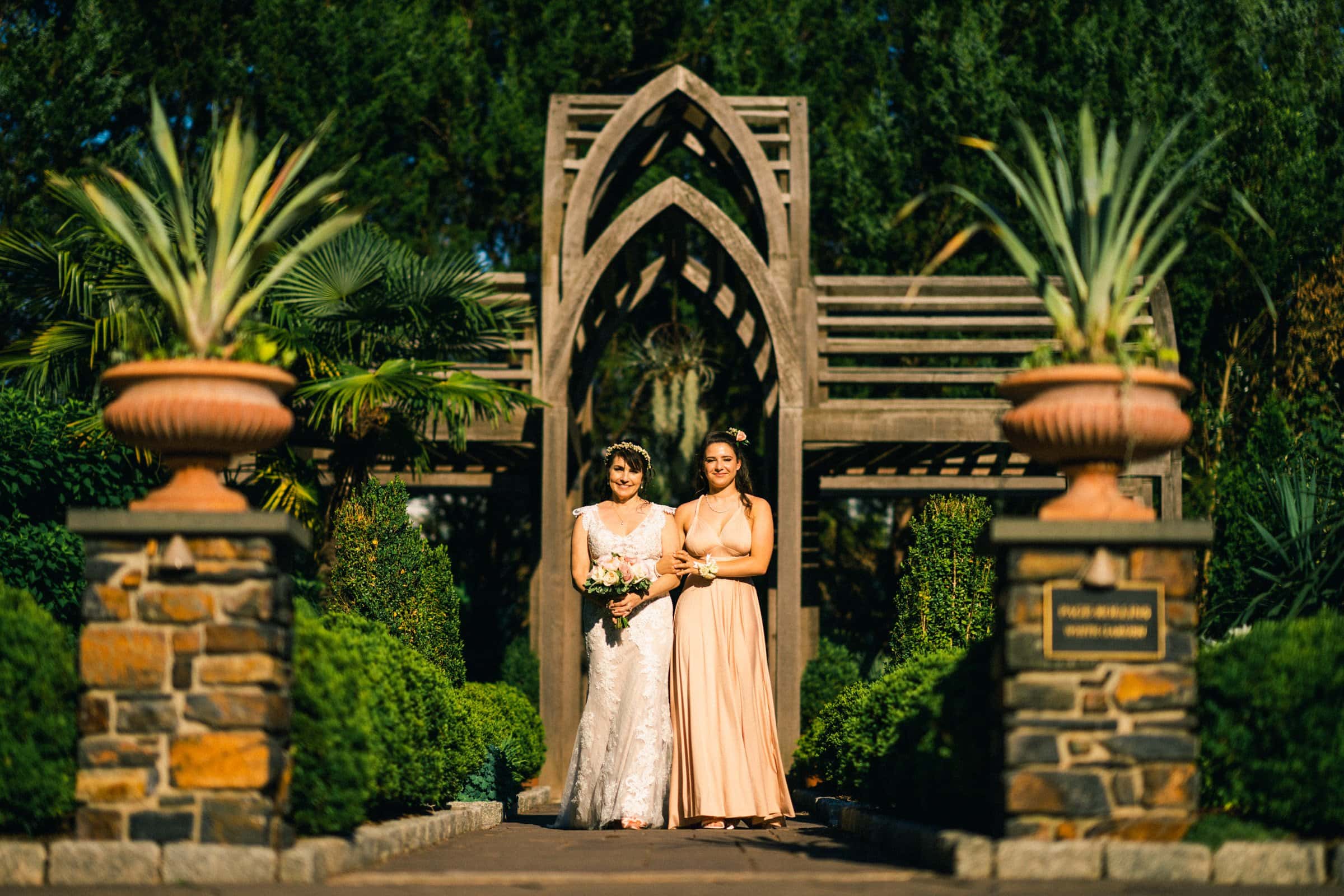 duke gardens wedding photography - ann & tim A bride in a white gown and a woman in a peach dress walk side by side down the garden path at Duke Gardens, lined with stone columns and large planters. An arched wooden structure stands in the background, surrounded by lush greenery, capturing an enchanting wedding photography moment.