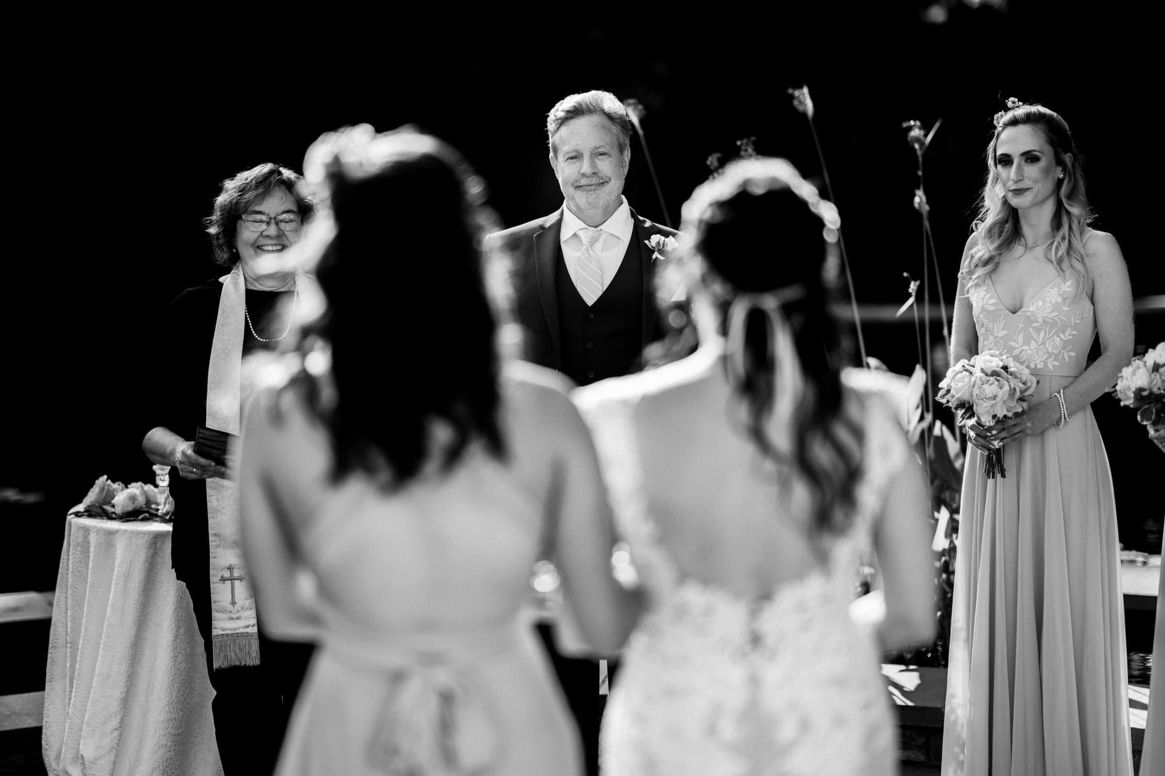 duke gardens wedding photography - ann & tim A black and white photo captures the essence of a Duke Gardens wedding ceremony. The groom stands beside the officiant, smiling, as two women with their backs to the camera look on. A bridesmaid holding a bouquet stands to the right in this serene outdoor setting.