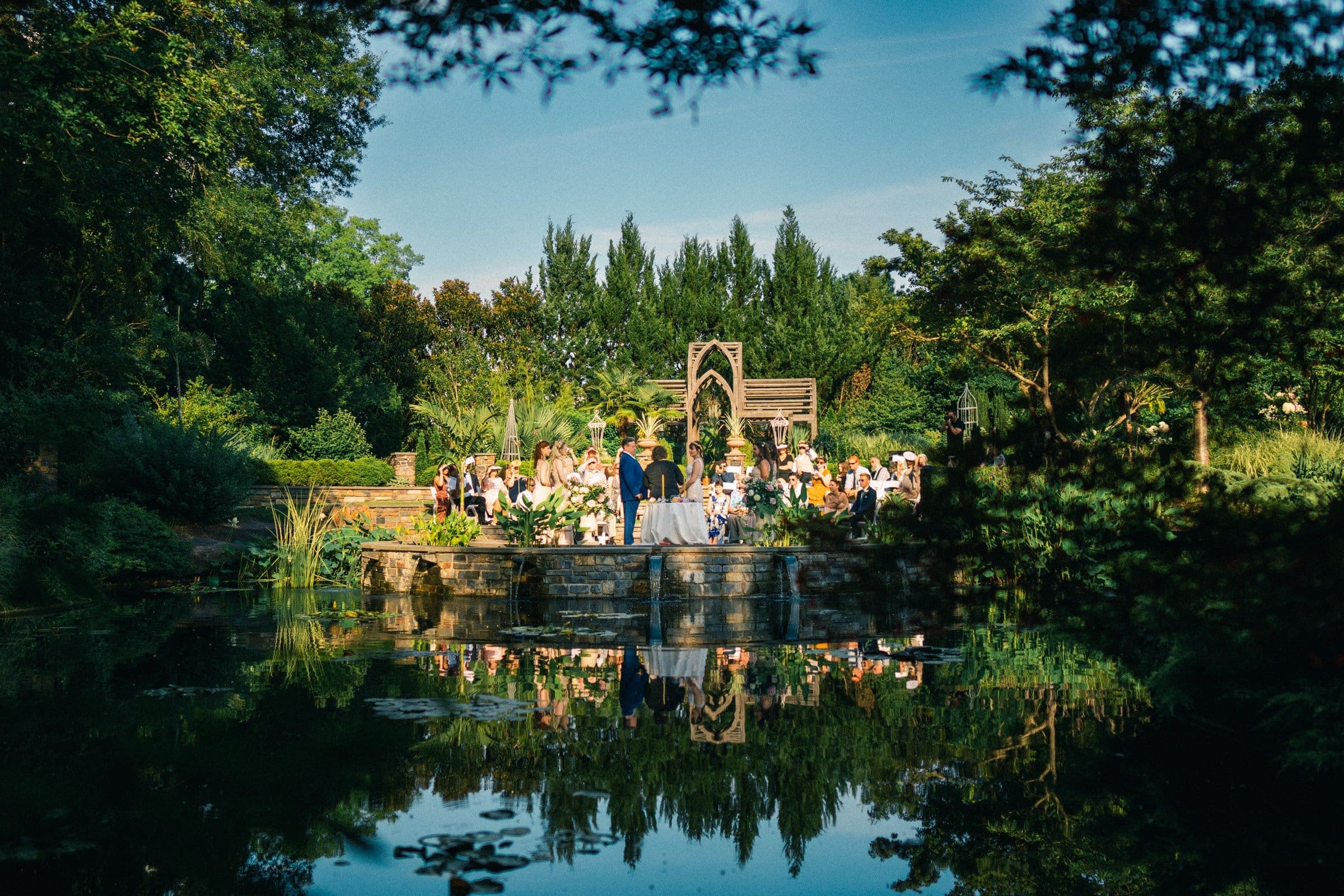duke gardens wedding photography - ann & tim A scenic outdoor wedding ceremony unfolds by a pond surrounded by lush greenery at Duke Gardens. Guests face the couple at the altar beneath a wooden arch, perfectly reflected in the tranquil water. The blue sky and trees enhance the serenity, ideal for capturing magical wedding photography.