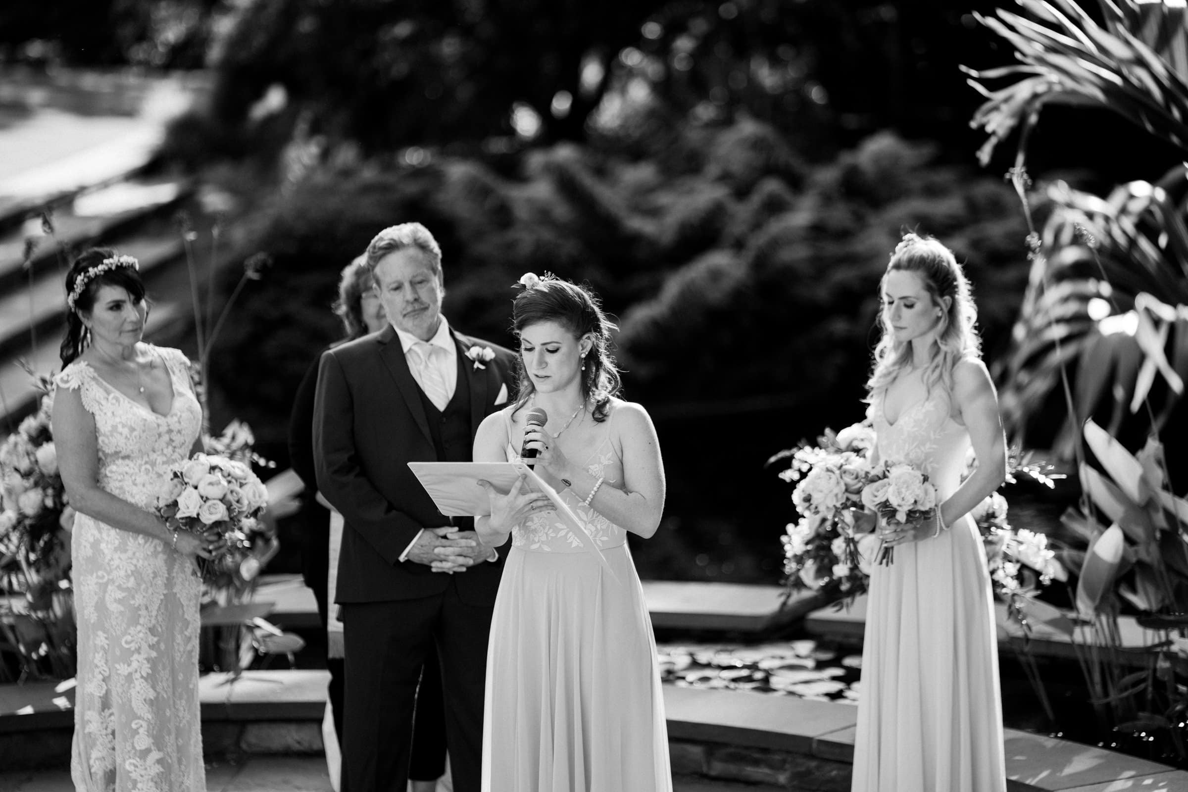 duke gardens wedding photography - ann & tim A black-and-white photo captures a Duke Gardens wedding ceremony. A woman stands in the center reading from a paper, flanked by a man and two women holding bouquets, while lush greenery and a water feature provide a serene backdrop.