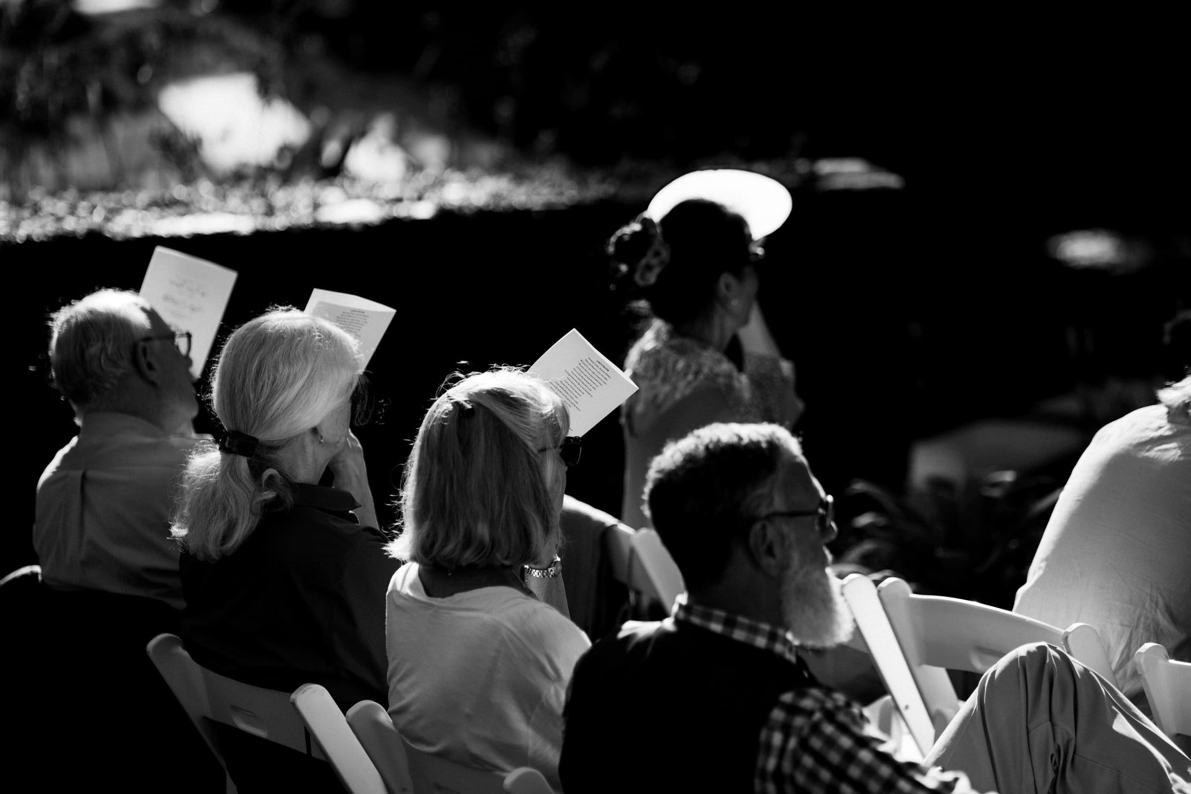 duke gardens wedding photography - ann & tim In a black and white image capturing the essence of Duke Gardens wedding photography, people are seated outdoors, holding papers or programs. They face away from the camera, basking in sunlight with trees and lush foliage creating a serene backdrop.