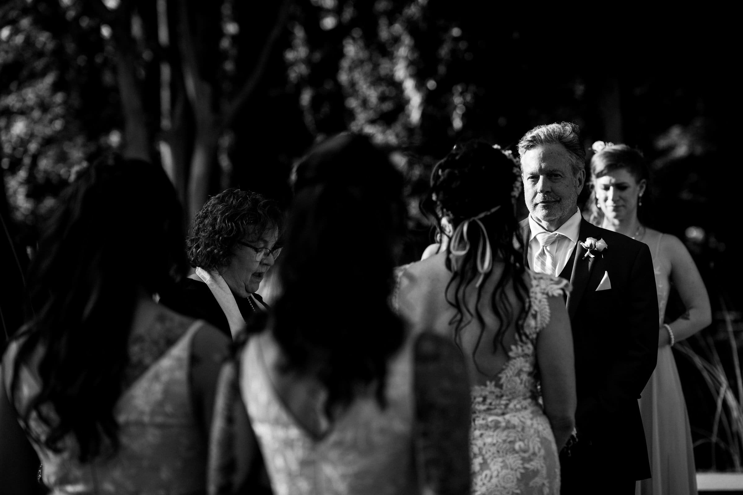 duke gardens wedding photography - ann & tim A black and white photo captures a Duke Gardens wedding ceremony. The groom, in a suit, faces the bride in her lace gown. They stand outdoors with an officiant and bridesmaids nearby. Towering trees and soft lighting enhance this intimate moment, immortalized by exquisite photography.