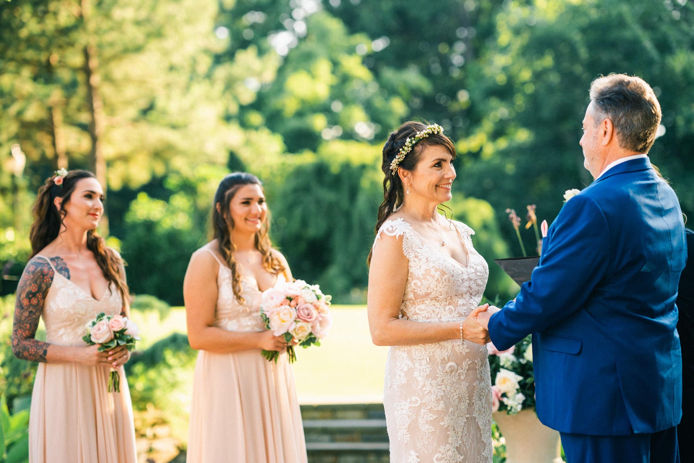 duke gardens wedding photography - ann & tim A bride and groom stand facing each other, holding hands during an outdoor ceremony at Duke Gardens. The bride is in a lacy dress, and the groom wears a blue suit. Two bridesmaids in pink dresses hold bouquets as they watch the couple amid the lush green backdrop, capturing perfect wedding photography.