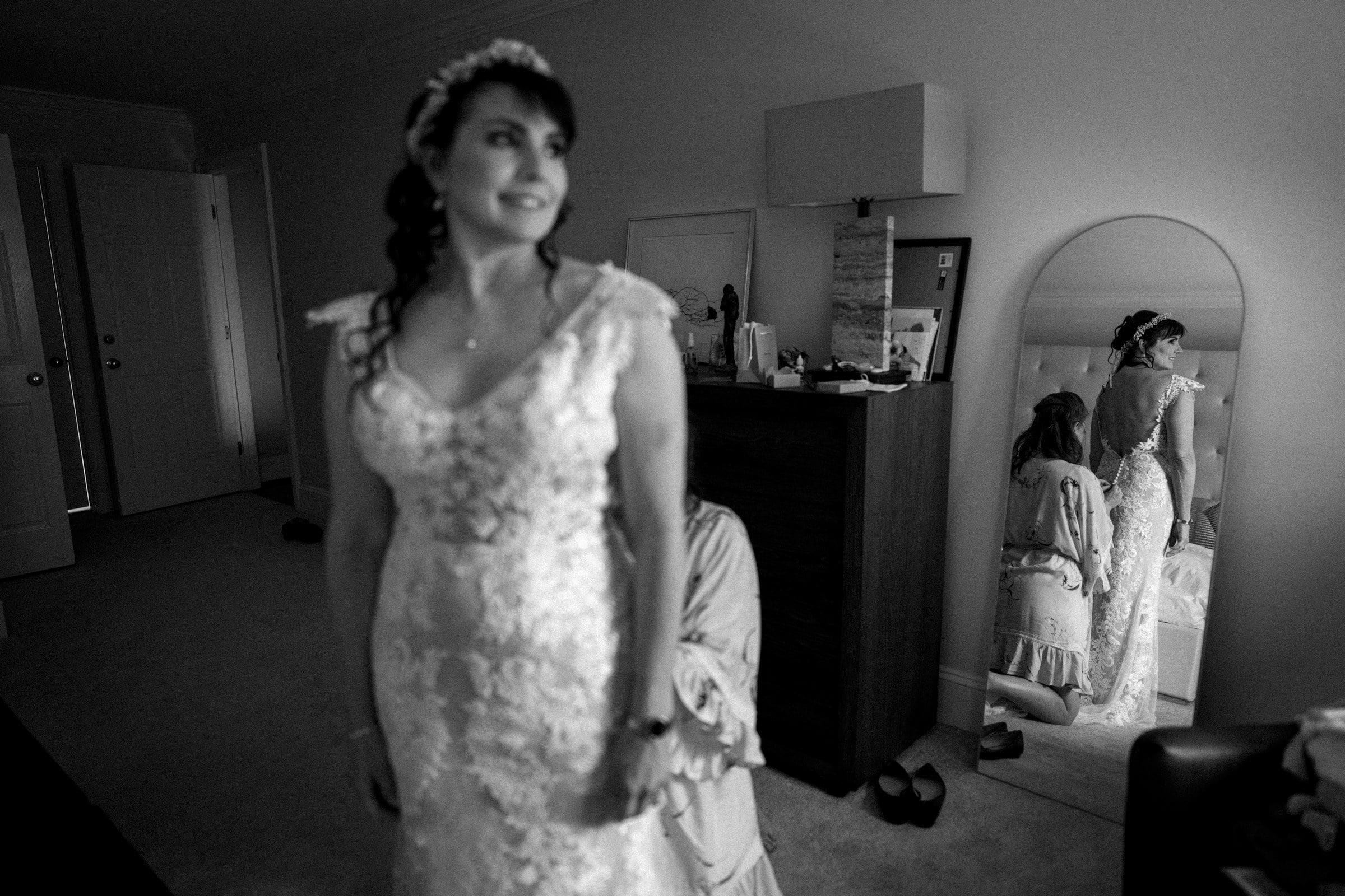 duke gardens wedding photography - ann & tim A bride in a lace dress stands gracefully in a softly lit room at Duke Gardens, her reflection captured perfectly in the mirror. Another person gently adjusts the dress from behind, with a dresser and a closed door accentuating the intimate wedding photography setting.