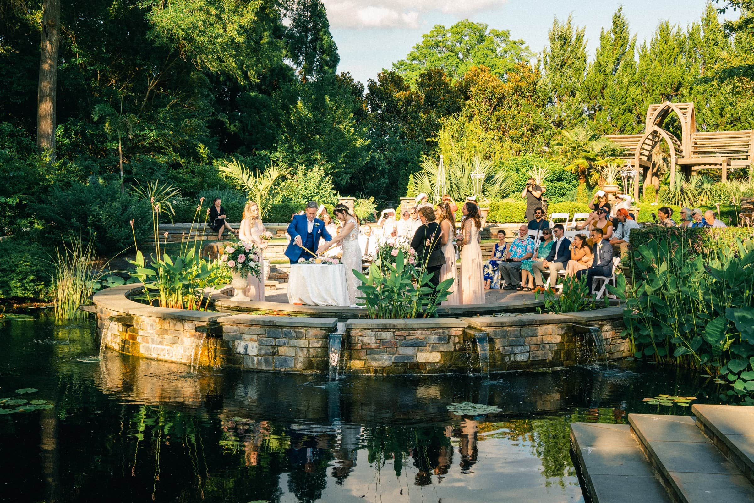 duke gardens wedding photography - ann & tim A wedding ceremony unfolds in a lush garden setting, perfect for Duke Gardens wedding photography. Guests gather around a stone platform near a serene pond, with trees and greenery enveloping the scene under a sunny sky.