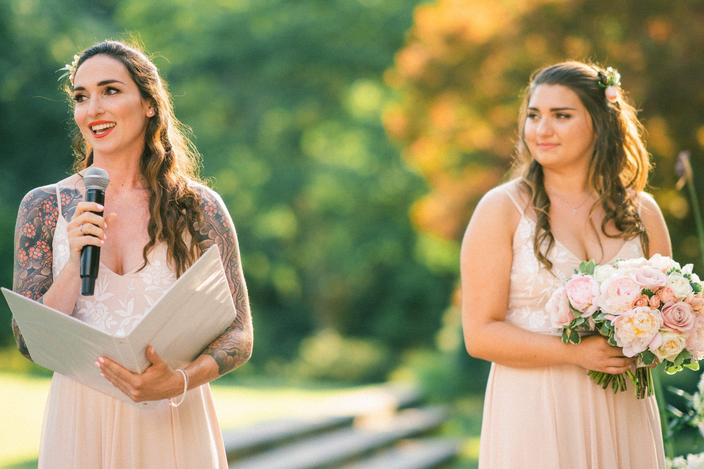 duke gardens wedding photography - ann & tim Two women in light dresses stand outdoors in what looks like a scene from Duke Gardens wedding photography. One, holding a microphone and papers, appears to be speaking while the other holds a bouquet of flowers. The lush greenery and sunlight filtering through the trees create a magical backdrop.