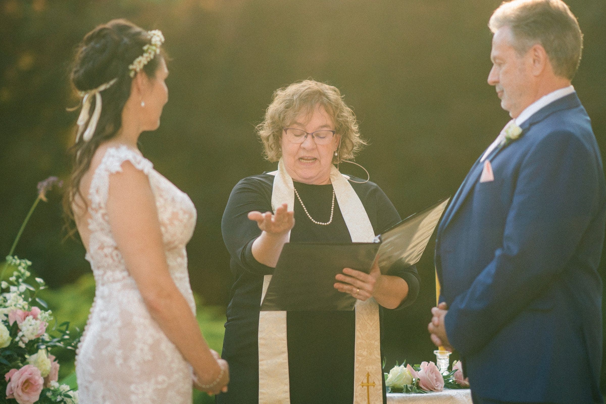 duke gardens wedding photography - ann & tim A woman in a white lace dress and a man in a blue suit stand before an officiant with curly hair, wearing a black dress and white stole, holding a book. They are outdoors at Duke Gardens, surrounded by greenery and flowers, as wedding photography captures the enchanting ceremony.