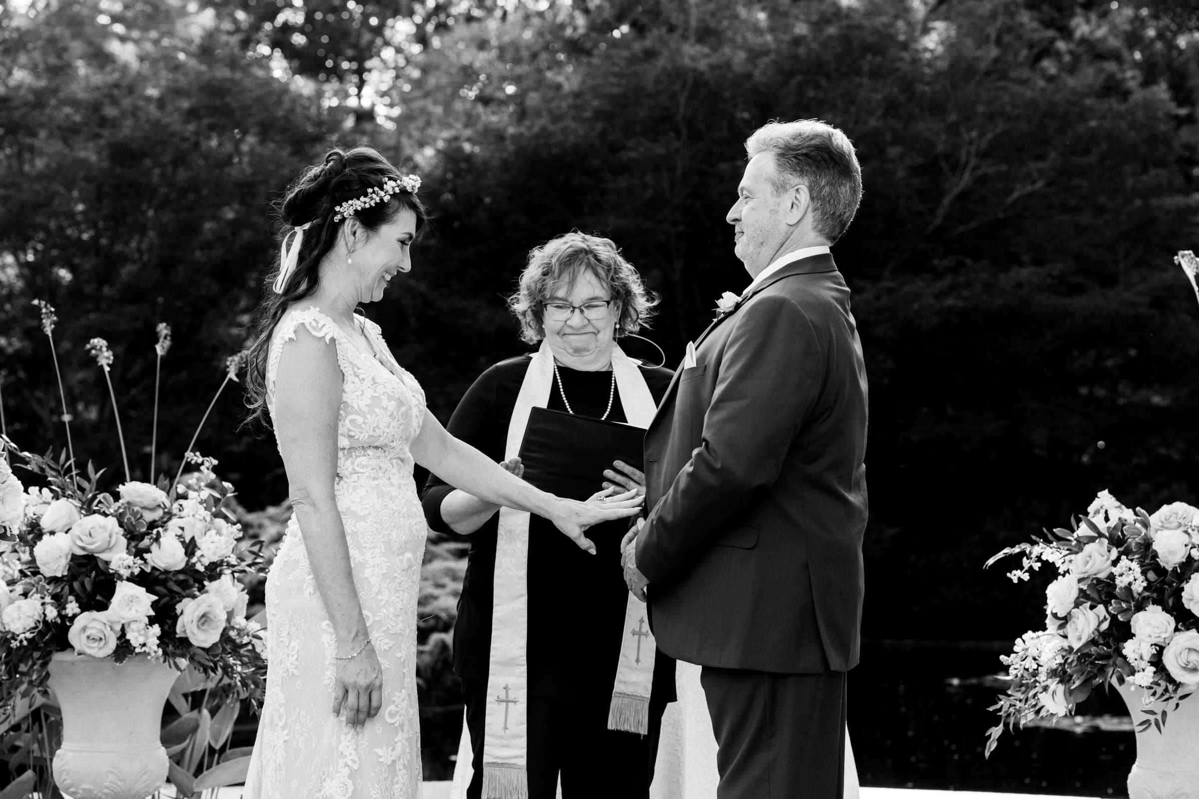 duke gardens wedding photography - ann & tim A black and white photo captures a Duke Gardens wedding ceremony. The bride, adorned in a lace dress, and the groom in a suit face each other, holding hands. An officiant stands between them, smiling, with trees and exquisite flower arrangements setting the perfect backdrop.