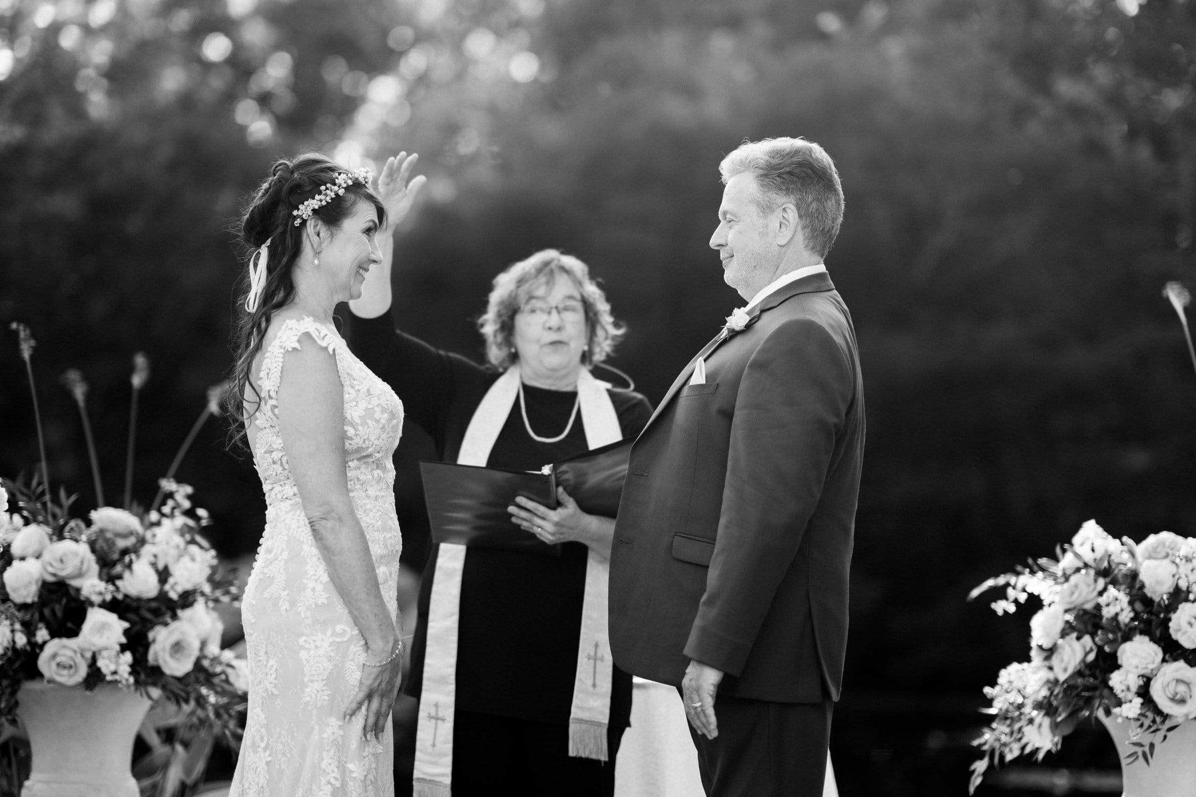 duke gardens wedding photography - ann & tim A bride and groom stand facing each other during an outdoor ceremony at Duke Gardens. A wedding officiant stands between them, holding a book and raising a hand. Elegant floral arrangements decorate the scene in this timeless black-and-white wedding photography masterpiece.