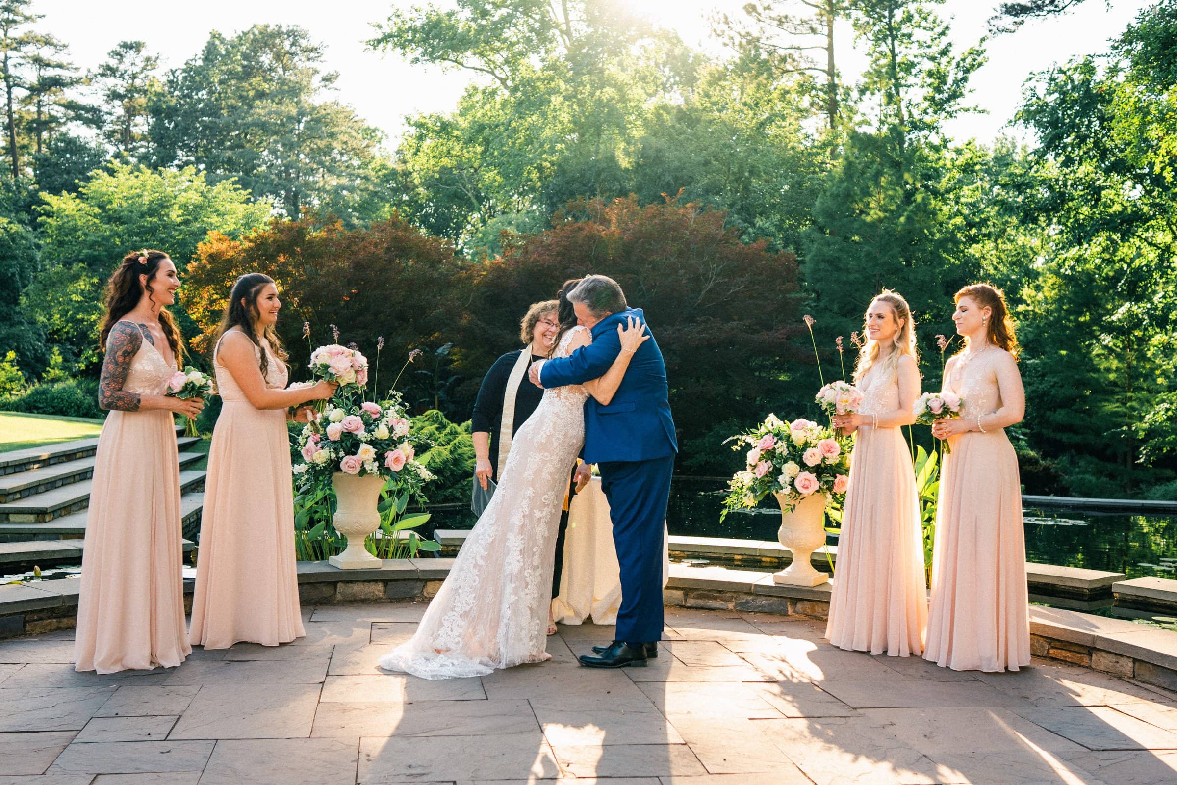duke gardens wedding photography - ann & tim A bride and groom share a kiss during an outdoor ceremony at Duke Gardens, surrounded by four bridesmaids in pink dresses. The officiant stands behind them amidst lush greenery and floral arrangements, capturing the perfect moment for wedding photography.