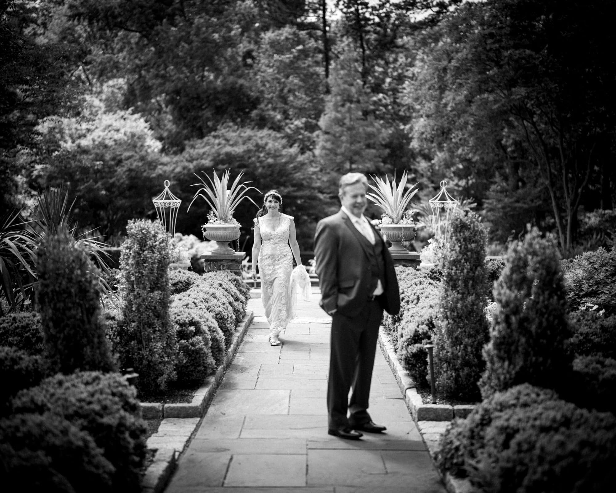 duke gardens wedding photography - ann & tim A black and white photo of a bride in a lace dress walking toward her groom in a suit on a garden path at Duke Gardens. Tall planters and decorative lanterns line the walkway, capturing an elegant outdoor setting perfect for wedding photography.