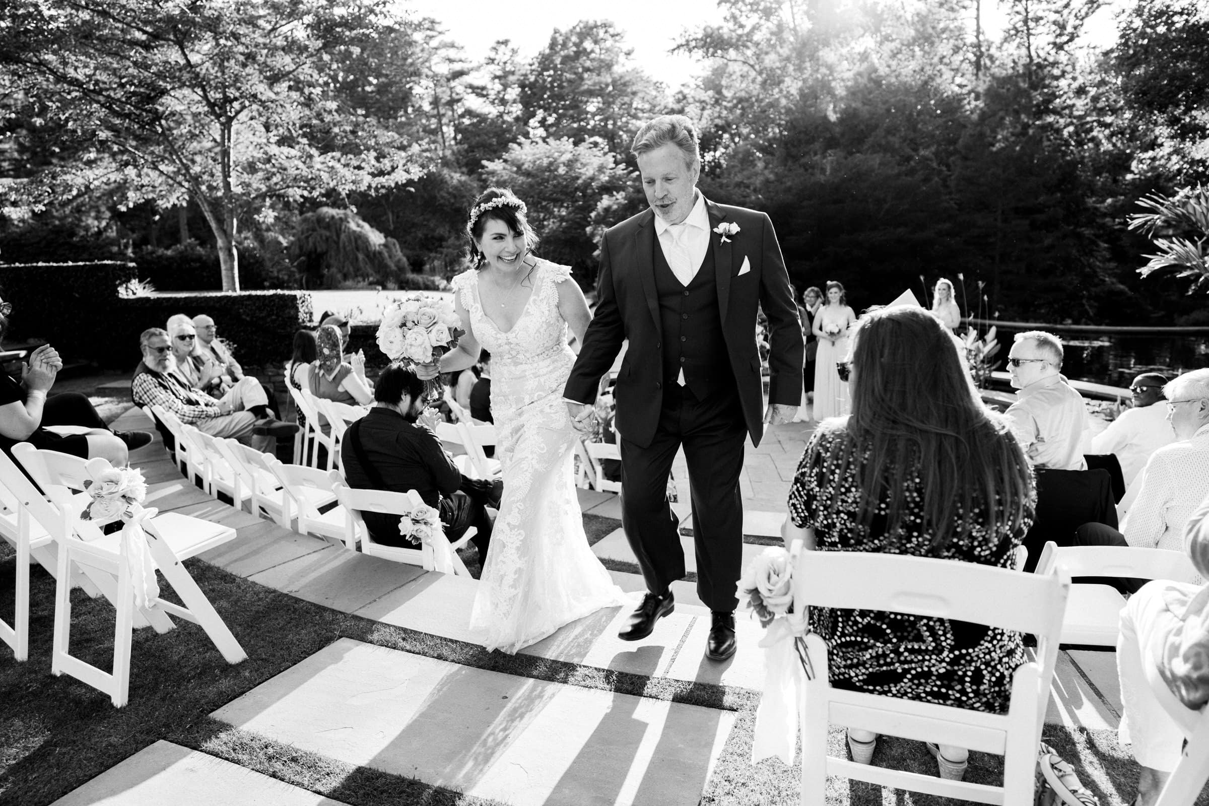 duke gardens wedding photography - ann & tim A newlywed couple walks joyfully down an outdoor aisle at Duke Gardens, with the bride in a lace gown and bouquet, and the groom in a suit. Guests in white chairs watch them, all beautifully captured by wedding photography amidst lush greenery and sunlight.