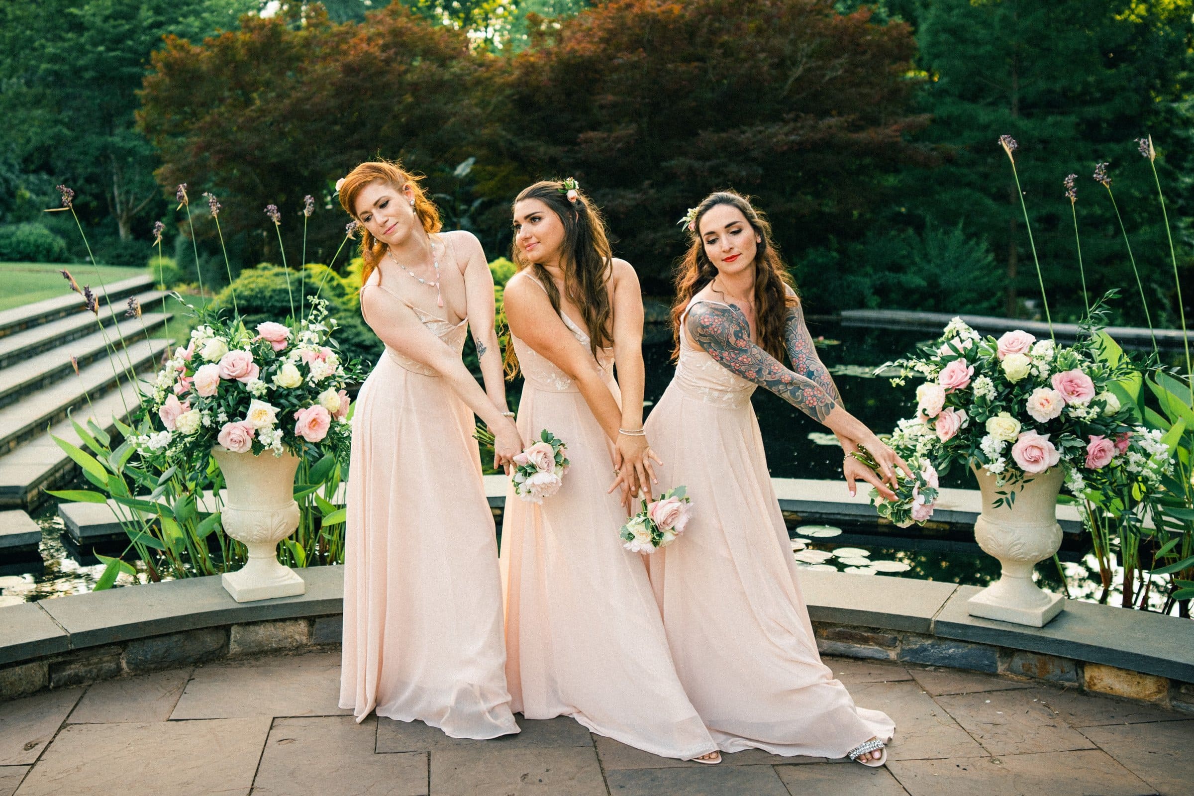 duke gardens wedding photography - ann & tim Three women in light pink dresses pose playfully in front of a pond with floral arrangements at Duke Gardens. They hold small bouquets and smile, with greenery and stone steps in the background, creating a serene, elegant wedding photography scene.