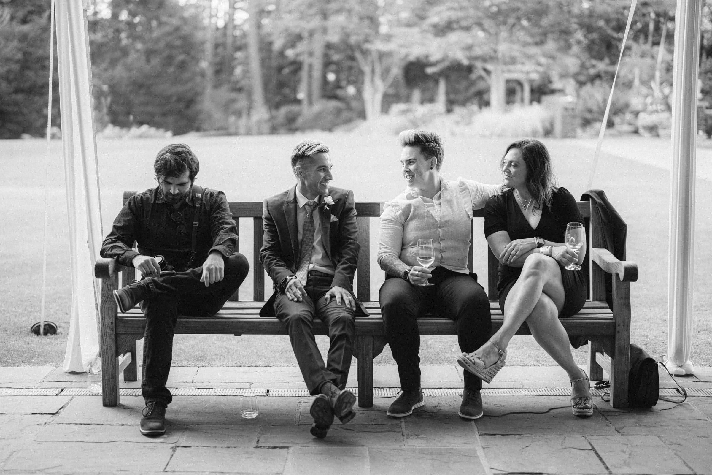 duke gardens wedding photography - ann & tim A black-and-white photo captures the essence of Duke Gardens wedding photography: four people on a bench, three men and a woman in formal attire, holding wine glasses, lost in conversation and laughter amidst the garden's beauty.