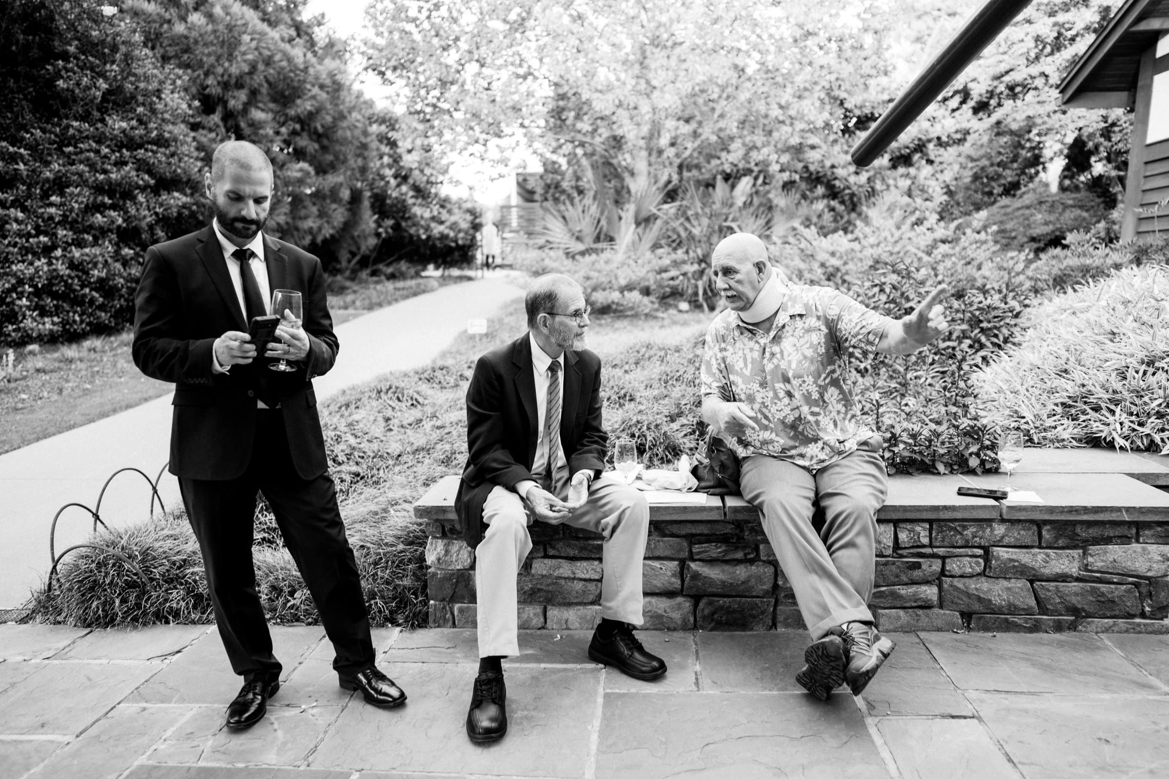 duke gardens wedding photography - ann & tim Three men are outdoors near a stone bench, captured in timeless black and white. One stands, looking at his phone, while the other two sit engaged in conversation. The setting evokes the serene elegance typical of Duke Gardens wedding photography, with trees and a pathway framing the scene.