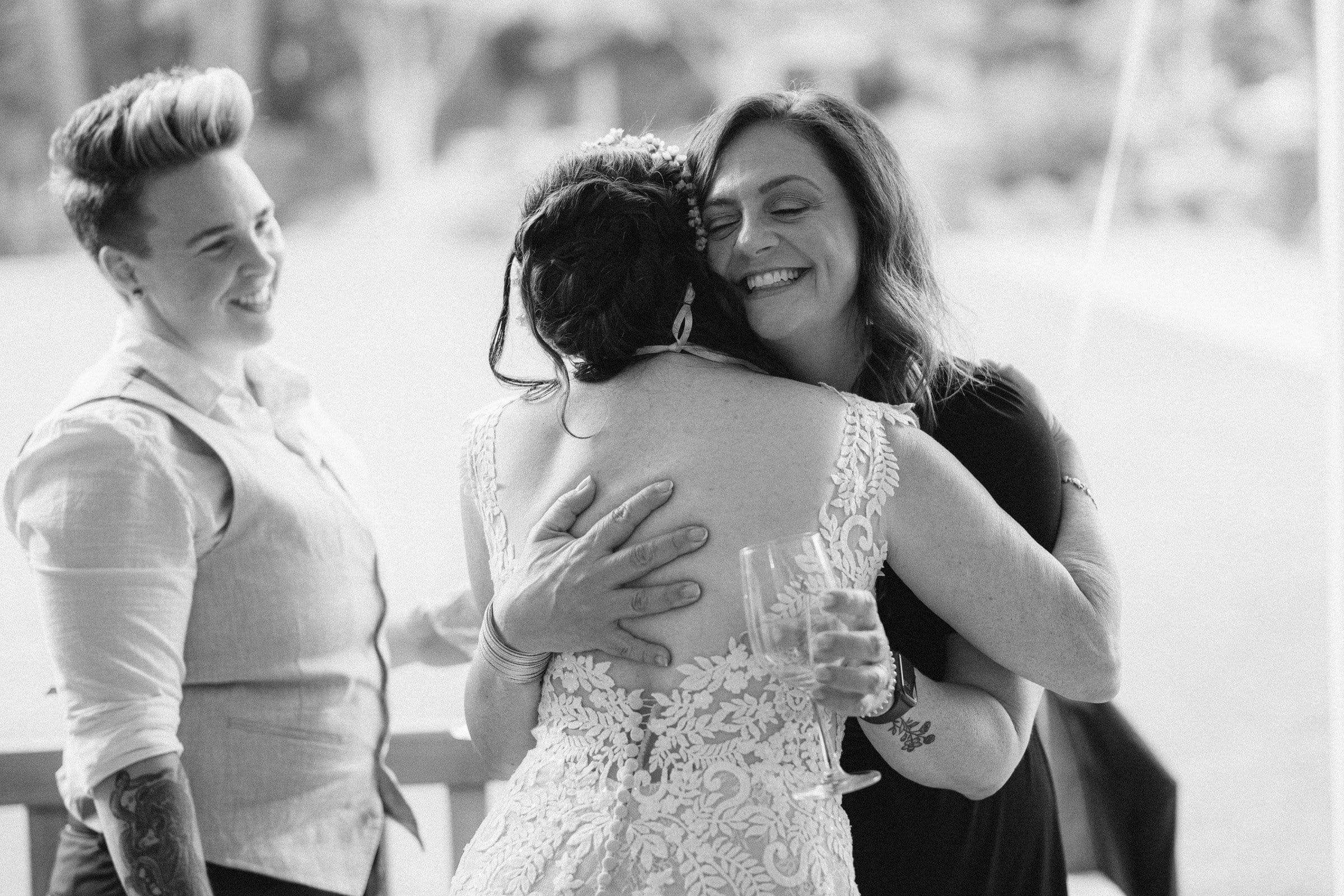 duke gardens wedding photography - ann & tim A black and white photo captures three people at a Duke Gardens wedding. One person in a lace dress is hugging another, who is smiling and holding a glass. Nearby, someone with short hair stands, also smiling. They're all enjoying the beautiful outdoor setting.