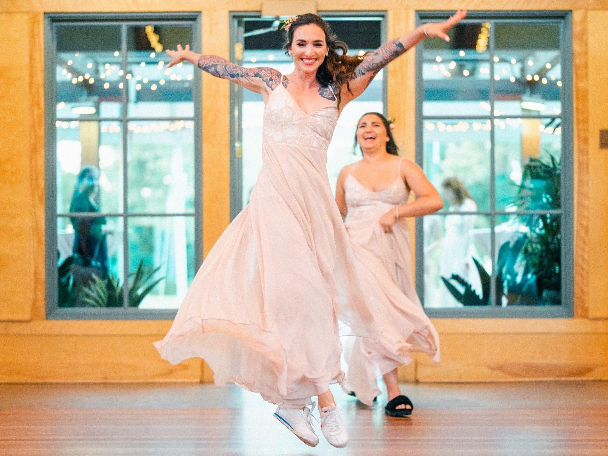 duke gardens wedding photography - ann & tim A woman in a light pink dress jumps with joy, her sneakers visible, as another woman in a similar dress smiles in the background. Indoors with large windows and string lights, this charming scene exudes the warmth of a Duke Gardens wedding photography session.