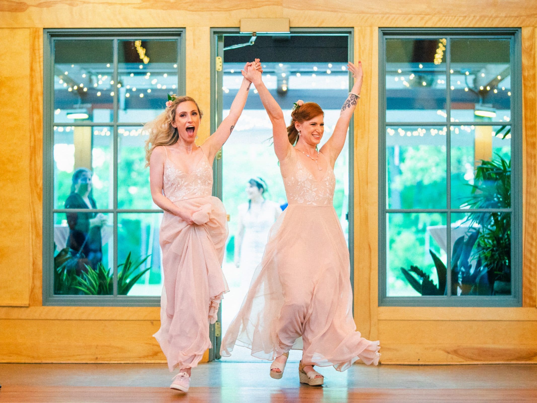 duke gardens wedding photography - ann & tim Two people in light pink dresses joyfully enter a room with their hands raised in celebration. They are smiling, and the background resembles Duke Gardens wedding photography, featuring large windows with greenery and string lights outside.