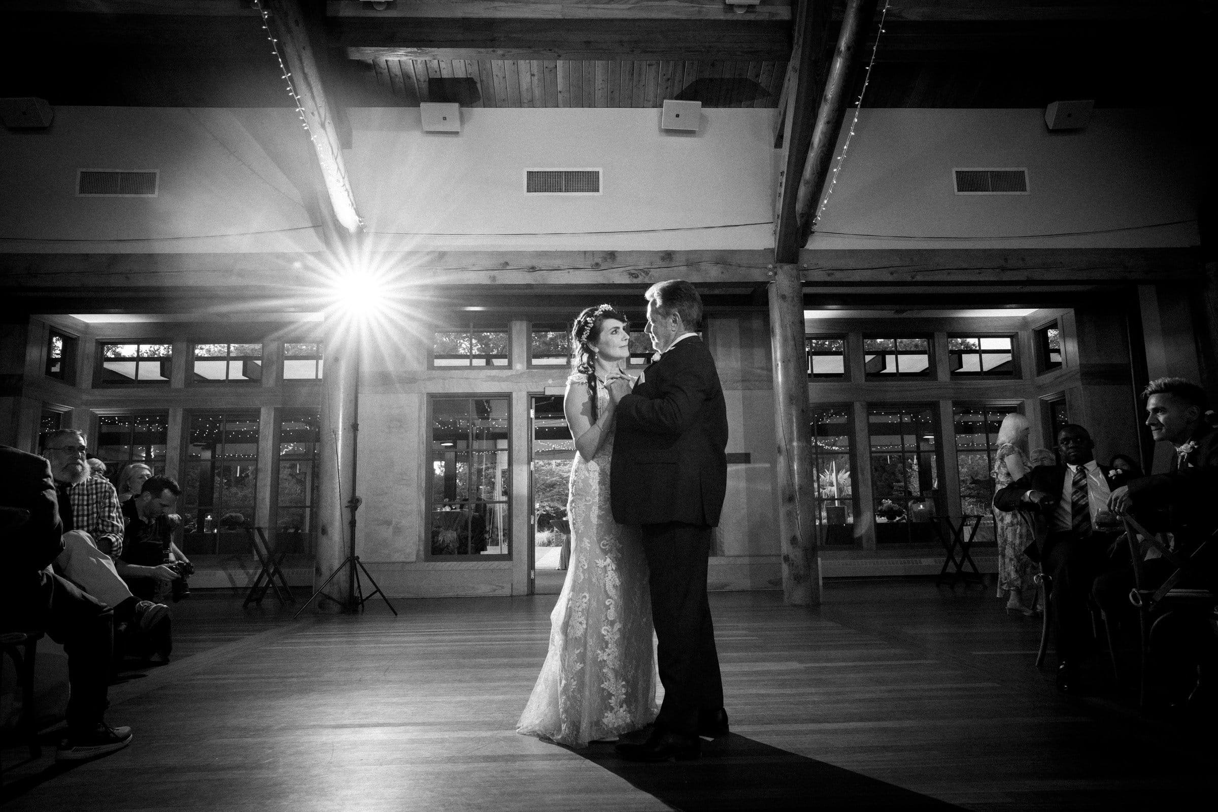duke gardens wedding photography - ann & tim A bride and groom share their first dance in a spacious reception hall with wooden beams and large windows. Captured in enchanting duke gardens wedding photography style, guests are seated around the room, and a bright light shines from the background, creating a dramatic atmosphere.