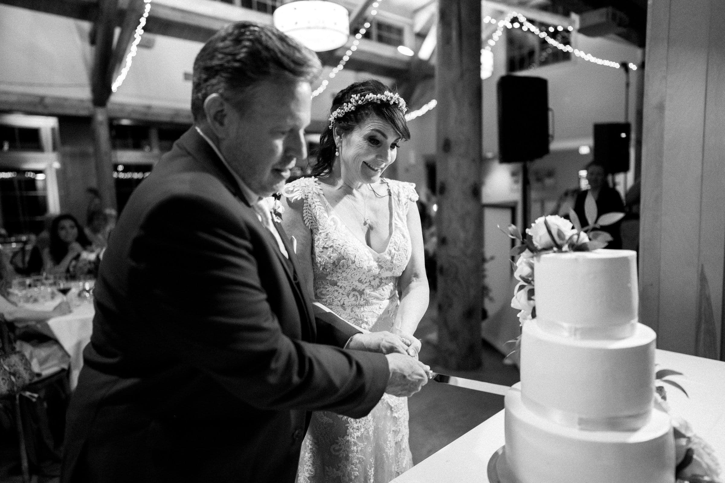 duke gardens wedding photography - ann & tim At a stunning Duke Gardens wedding reception, the bride and groom joyfully cut into a three-tiered cake. The venue glows with string lights, accentuating the bride's lace dress and headpiece as the groom stands beside her in a suit. Guests cheer from elegantly set tables in the background.
