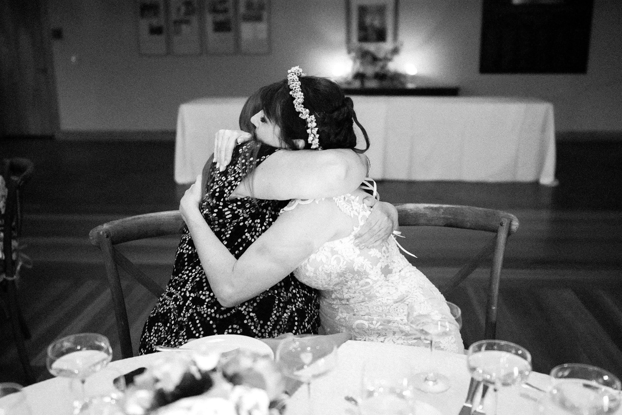 duke gardens wedding photography - ann & tim A bride in a lace dress and pearl headpiece hugs a guest at a wedding reception, captured beautifully in Duke Gardens wedding photography. Empty glasses and a floral centerpiece adorn the table in the softly lit room, with blurred decorations adding to the enchanting backdrop.