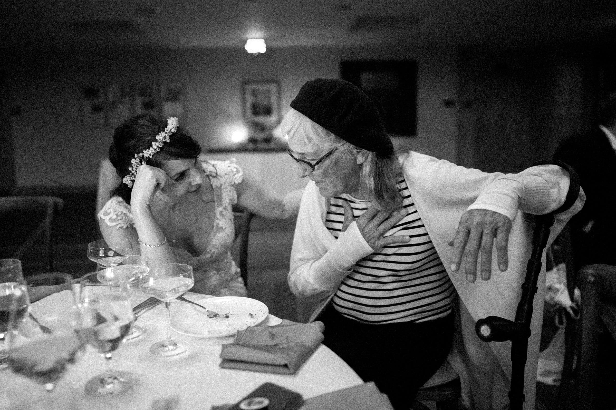 duke gardens wedding photography - ann & tim In the softly lit room, a bride in a white dress and floral headband leans in, smiling, to speak with an older person wearing glasses and a beret. Capturing this tender moment of their Duke Gardens wedding photography are plates and glasses on the table they share.