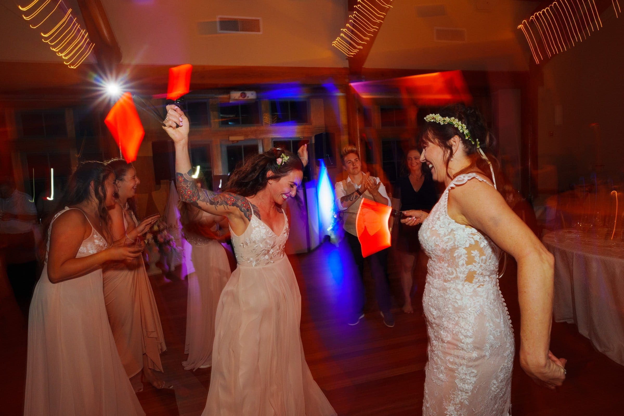 duke gardens wedding photography - ann & tim A group of people, dressed in formal attire, joyfully dancing at a Duke Gardens wedding reception. The room is softly lit with colorful lights in motion, creating a lively and celebratory atmosphere. Two women in wedding dresses are at the center of the scene—a perfect moment for wedding photography.