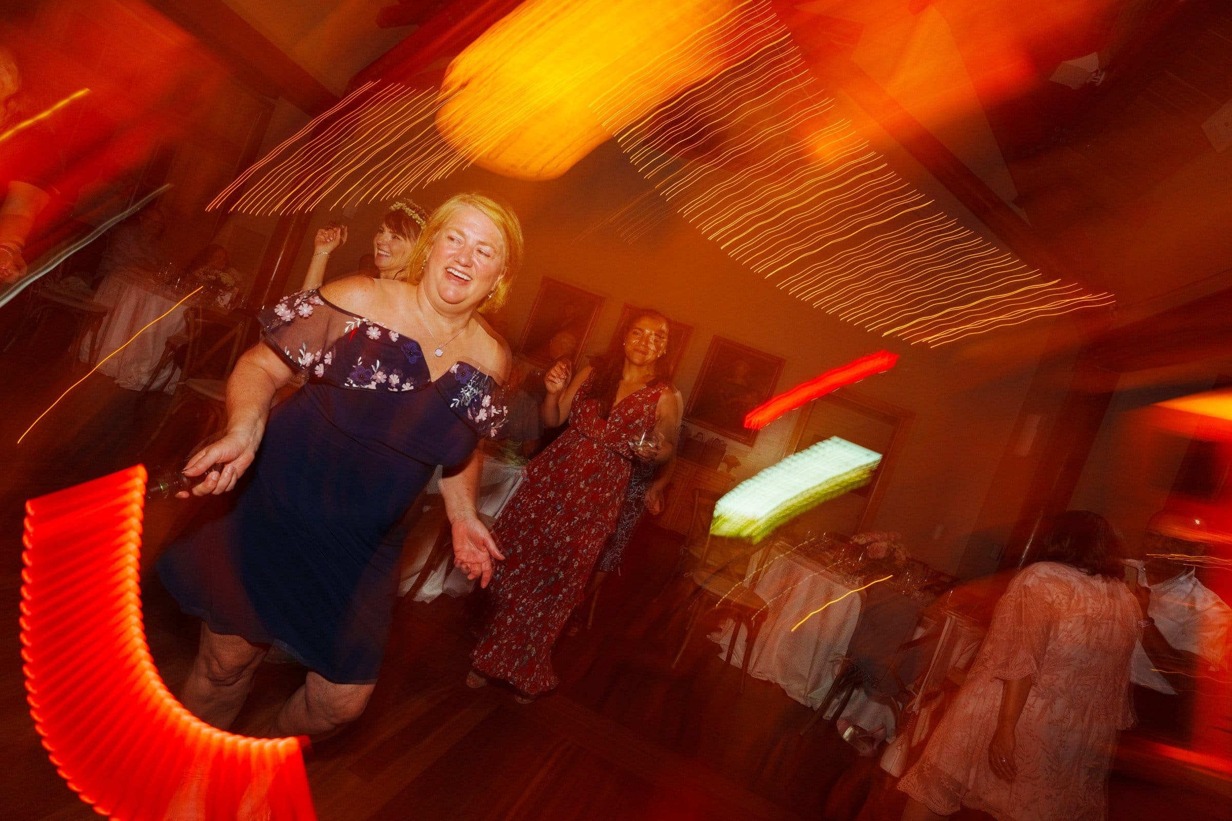 duke gardens wedding photography - ann & tim A joyful woman in a dark blue dress dances energetically in a room lit with warm, swirling lights. As others join her in the background, they create a lively and festive atmosphere reminiscent of Duke Gardens wedding photography.