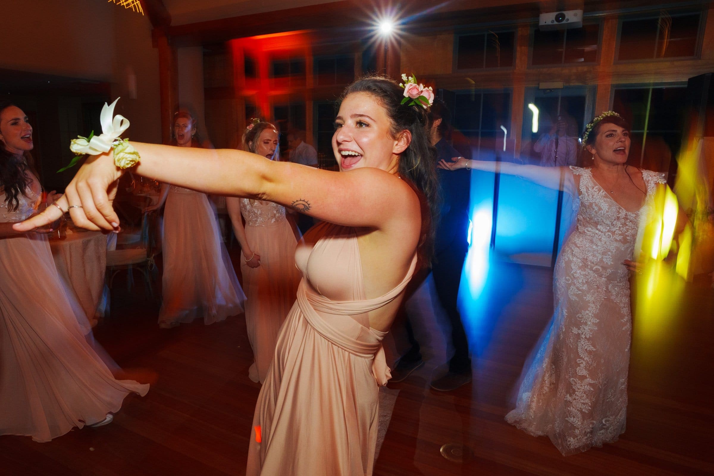 duke gardens wedding photography - ann & tim A joyful woman dancing at a wedding, wearing a light-colored gown and a flower corsage. Other guests, also in elegant dresses, dance in the background at Duke Gardens, with colorful lighting creating a festive atmosphere perfect for wedding photography.