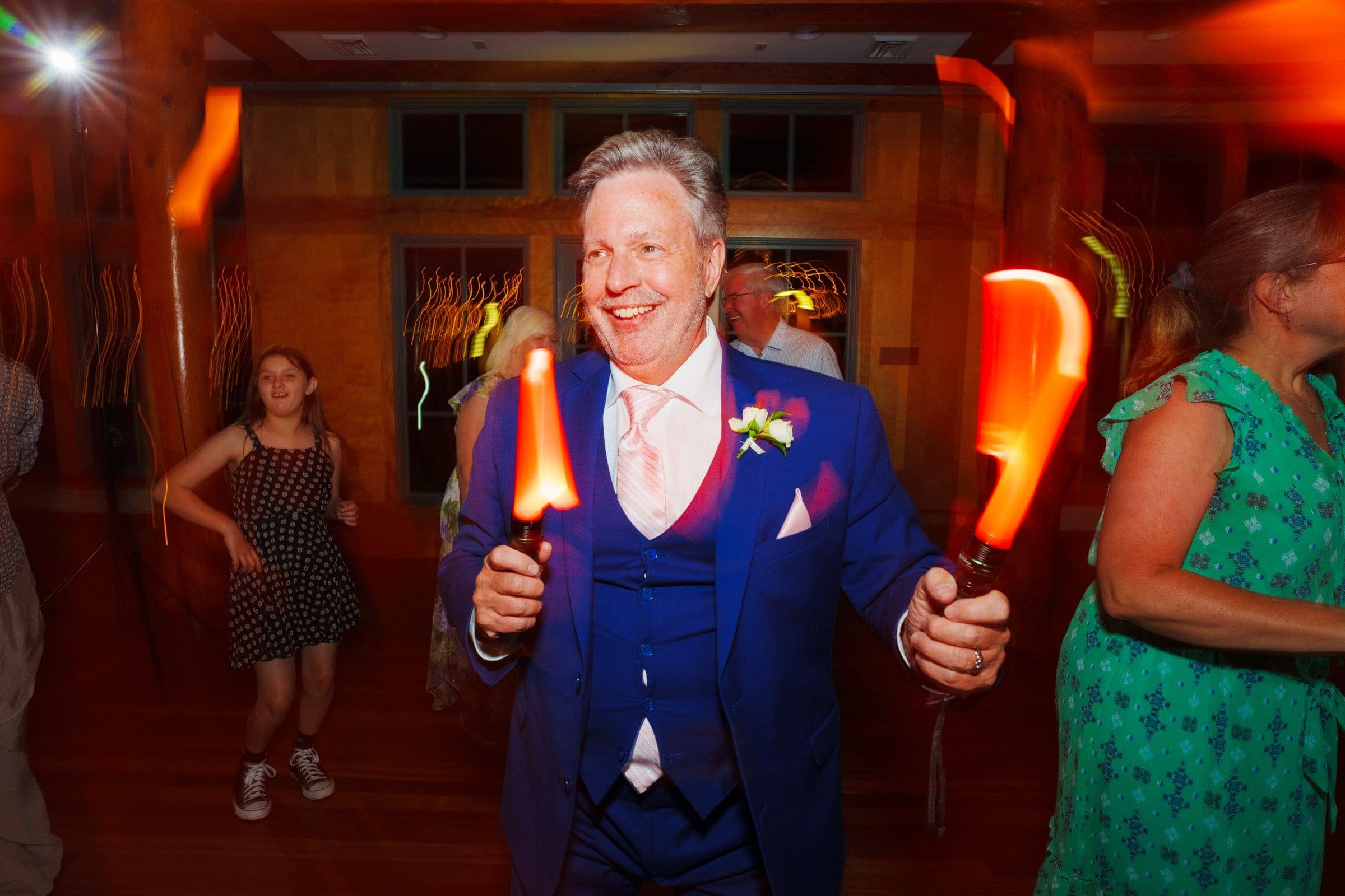 duke gardens wedding photography - ann & tim At a lively indoor event, reminiscent of Duke Gardens wedding photography, a man in a blue suit dances joyfully with light-up wands. Other people are blurred in the background, enhancing the dynamic and festive atmosphere.