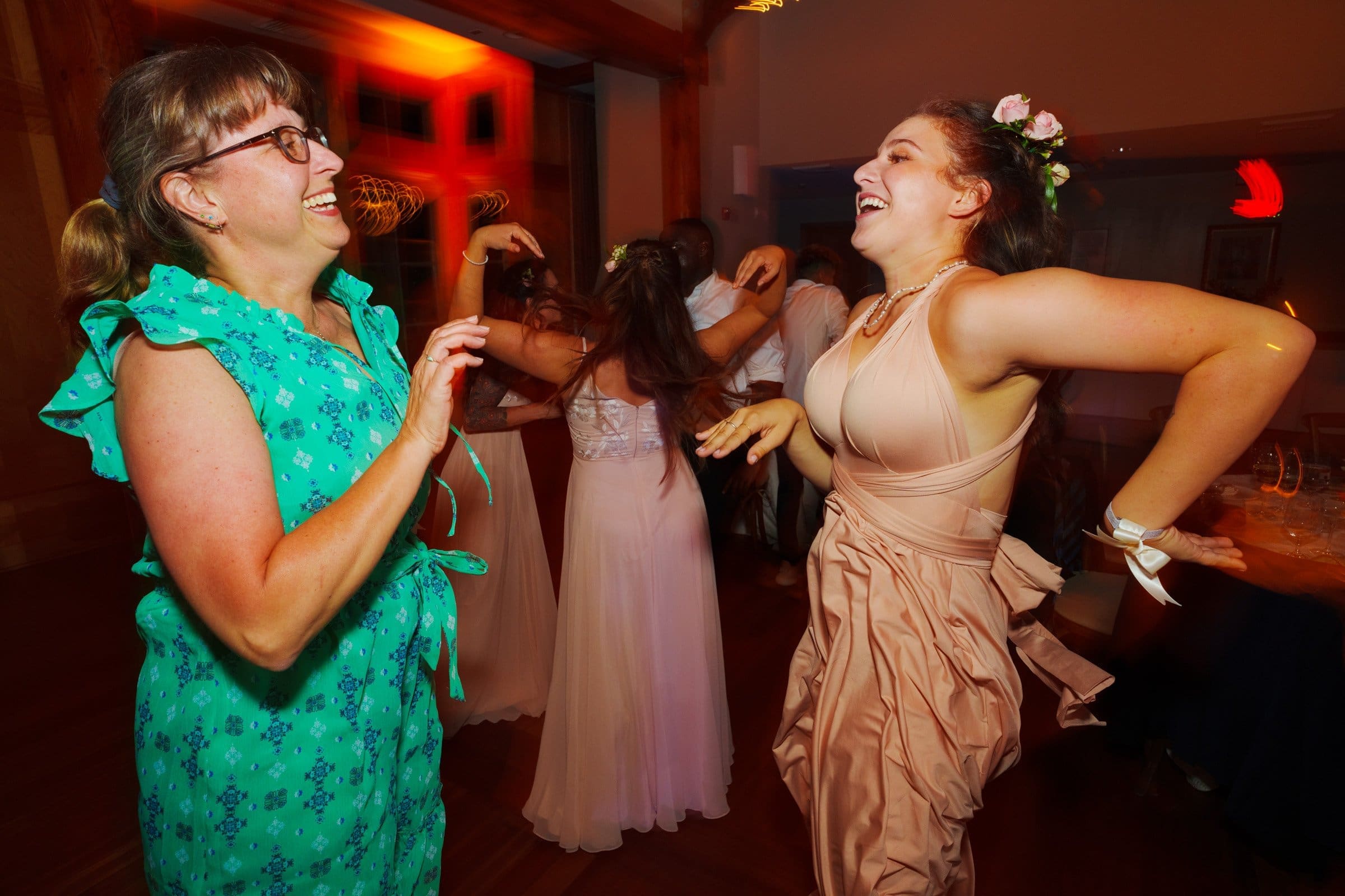 duke gardens wedding photography - ann & tim Two women joyfully dance at a lively indoor event, captured beautifully like a scene out of Duke Gardens wedding photography. The woman on the left wears glasses and a green floral dress, while the one on the right stuns in a pink dress and floral headpiece. The warmly lit background adds to the enchanting atmosphere.