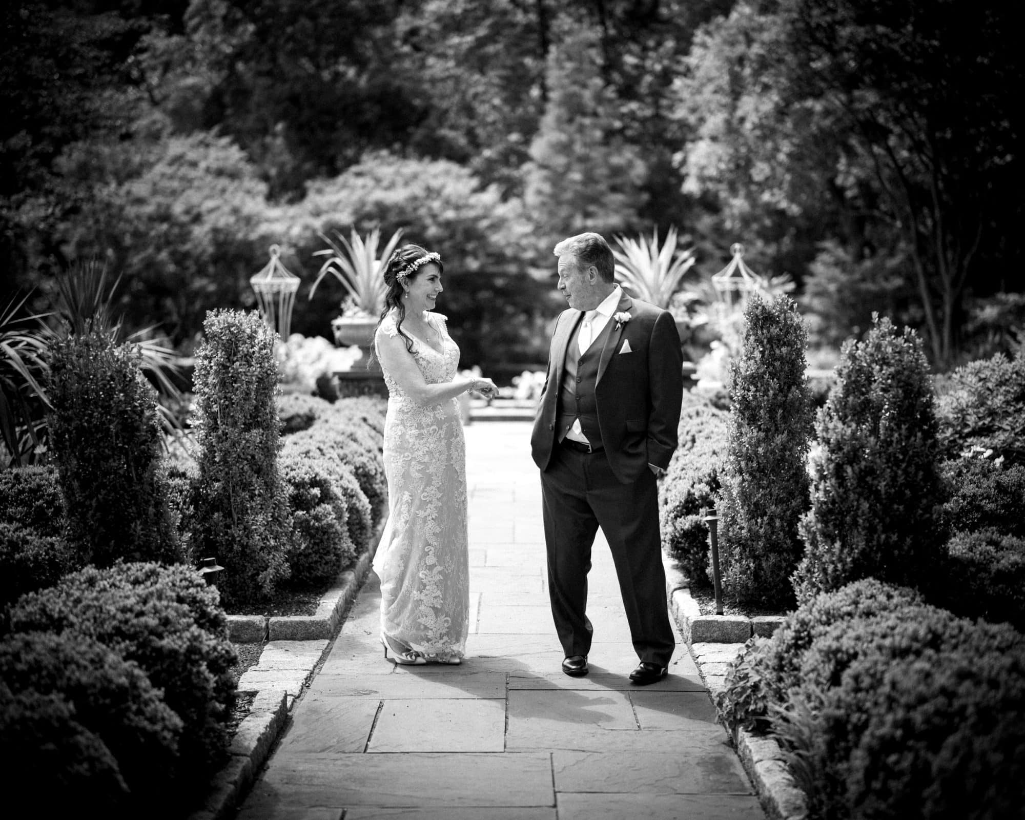 duke gardens wedding photography - ann & tim A bride and groom stroll along a garden path, enveloped by the elegant beauty of Duke Gardens. The serene scene is in black and white, capturing their formal attire as they gaze at each other with joyful smiles, embodying timeless wedding photography.