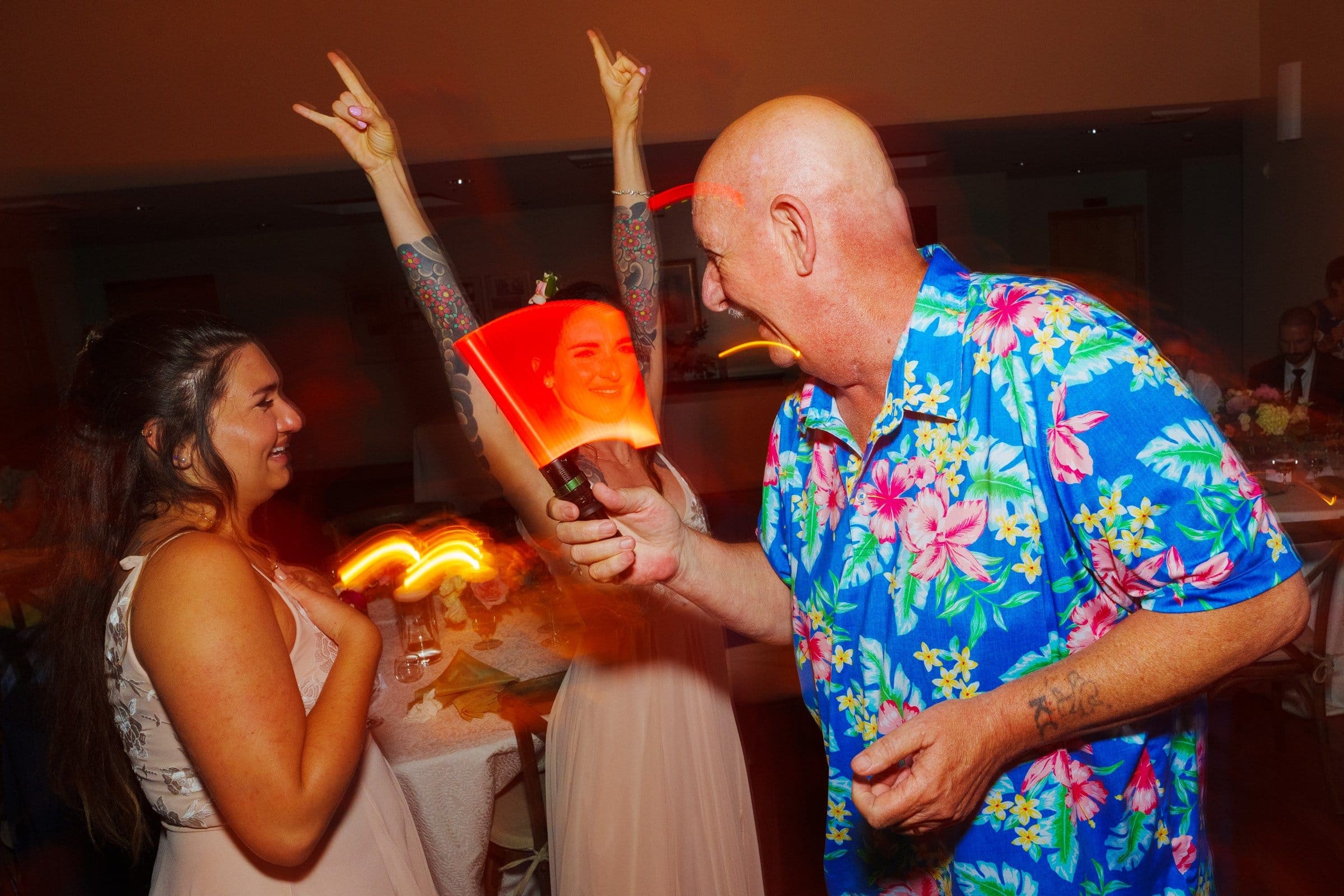 duke gardens wedding photography - ann & tim A joyful scene reminiscent of a Duke Gardens wedding photography session unfolds at the party, with three people dancing and smiling. A bald man in a colorful floral shirt holds a light stick, while two women, one with raised arms, enjoy the moment as blurred lights create a dynamic backdrop.