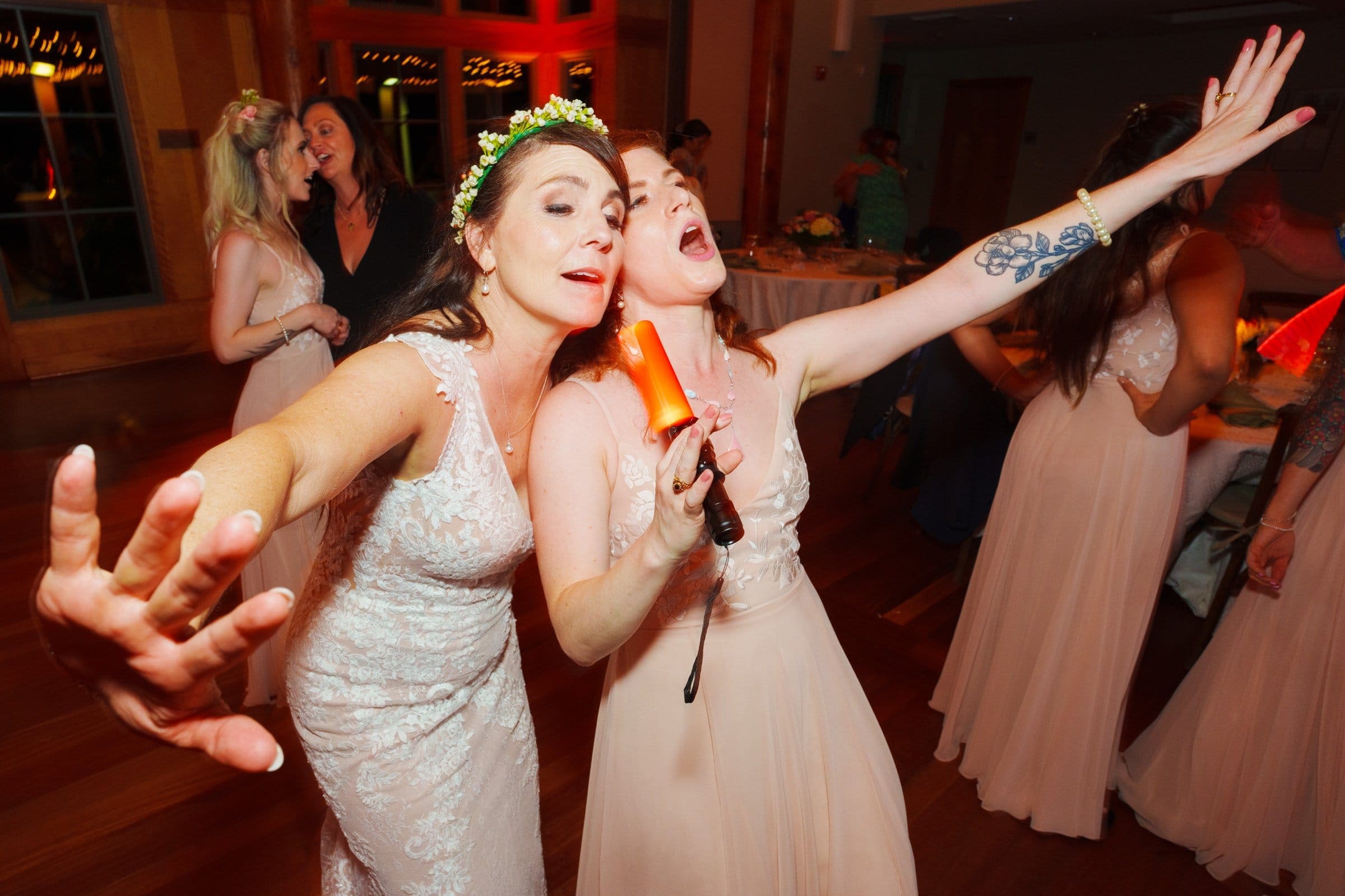 duke gardens wedding photography - ann & tim Two women in elegant dresses, one holding a microphone and singing passionately, the other with an arm extended. Both are joyful, wearing floral headpieces. Capturing this enchanting moment of music and dance at Duke Gardens evokes the essence of wedding photography.