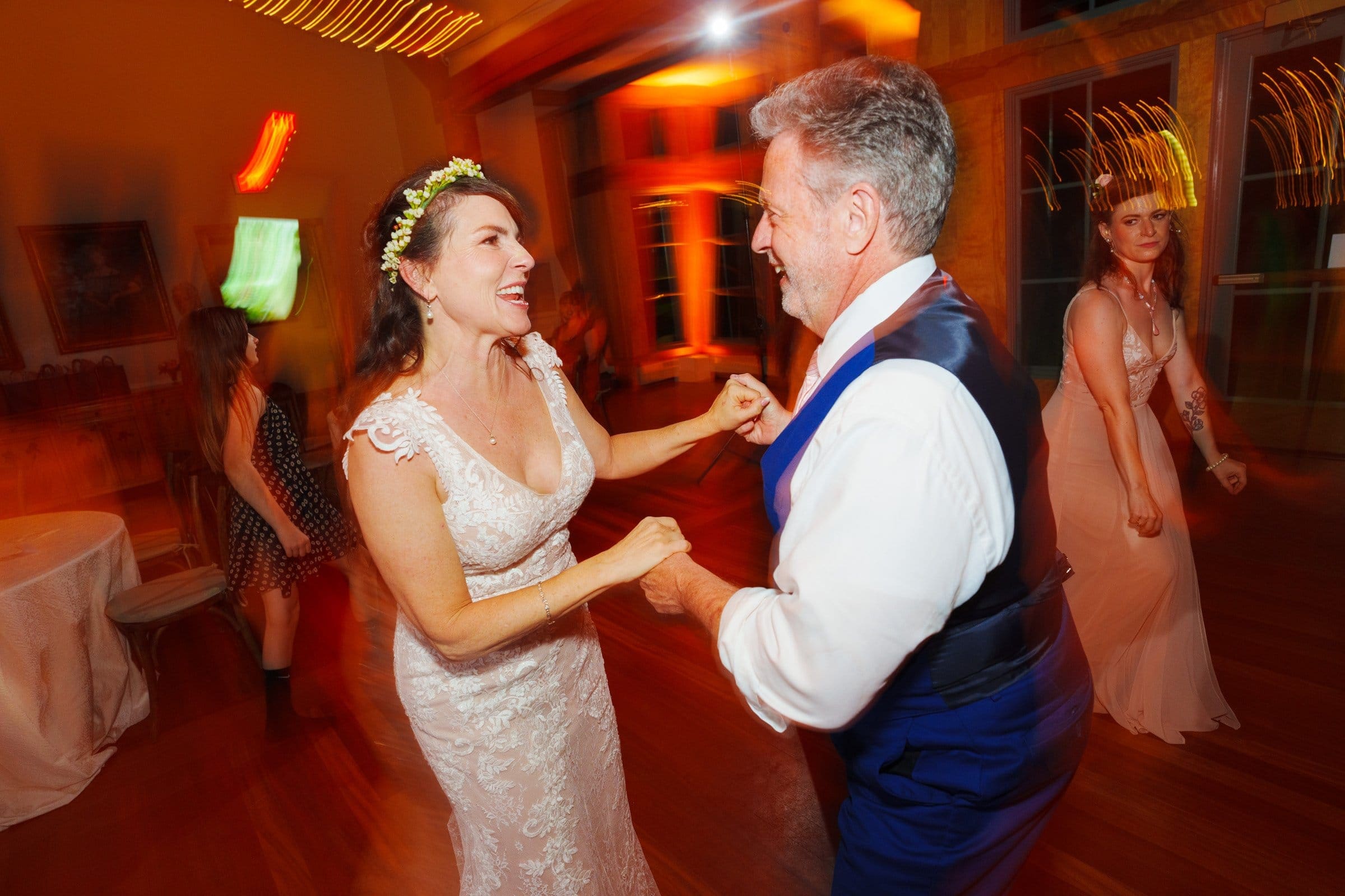duke gardens wedding photography - ann & tim At a charming indoor wedding reception reminiscent of Duke Gardens' elegance, a couple dances, smiling and holding hands. The woman, in a white dress with a flower crown, twirls gracefully with the man in a white shirt and blue vest. A bridesmaid and guests are visible in the warmly lit background.