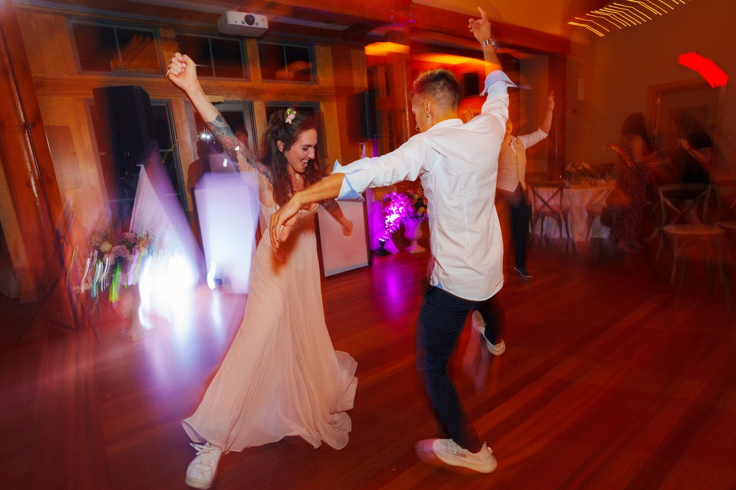 duke gardens wedding photography - ann & tim A woman in a flowing dress and a man in a white shirt and dark pants dance energetically on a wooden floor at Duke Gardens, with colorful lights and blurred motion capturing the lively essence of wedding photography.