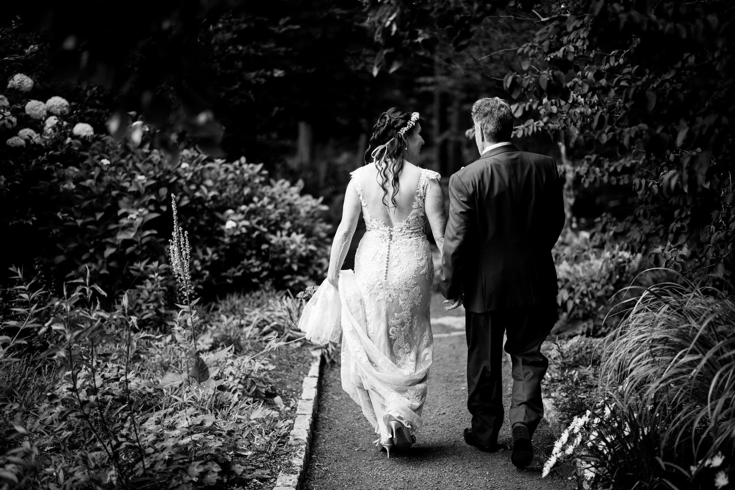duke gardens wedding photography - ann & tim In a timeless black and white photo, a couple walks hand in hand down a lush garden path, exuding elegance in their wedding attire—a lace gown for the bride and a suit for the groom. The serene atmosphere mirrors the charm of Duke Gardens wedding photography.