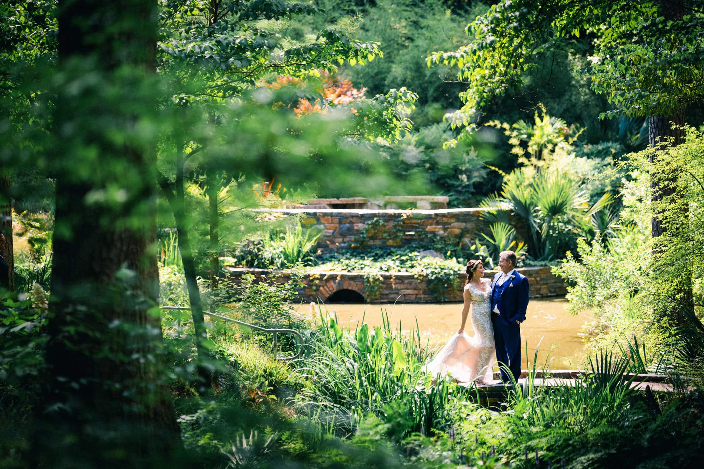 duke gardens wedding photography - ann & tim A couple in wedding attire stands by a pond amidst the lush greenery of Duke Gardens. The bride, in a flowing white dress, and the groom, in a blue suit, pose gracefully. This serene garden setting, complete with trees, bushes, and a stone bridge, is perfect for wedding photography.