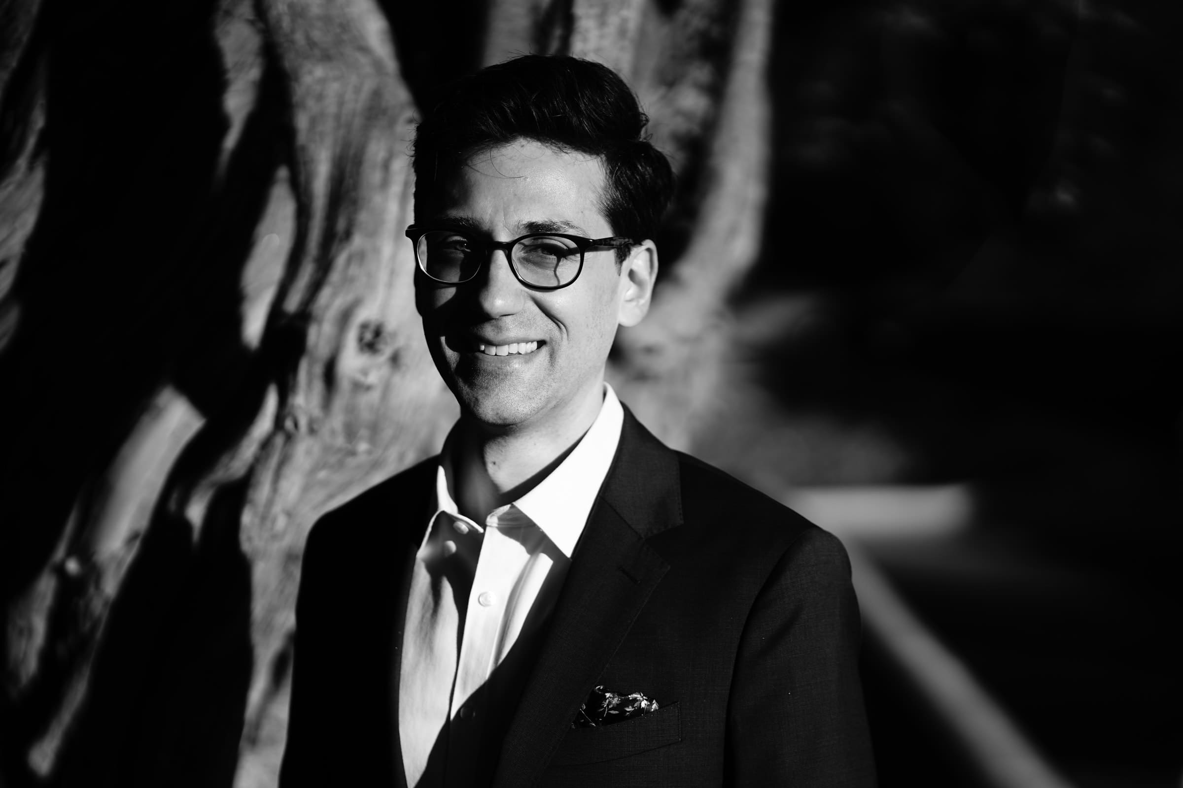 A black and white photo captures a person in a suit and glasses, smiling beneath the sprawling shadows of a textured tree. This outdoor scene radiates the timeless elegance often seen in Biltmore estate engagement photography.
