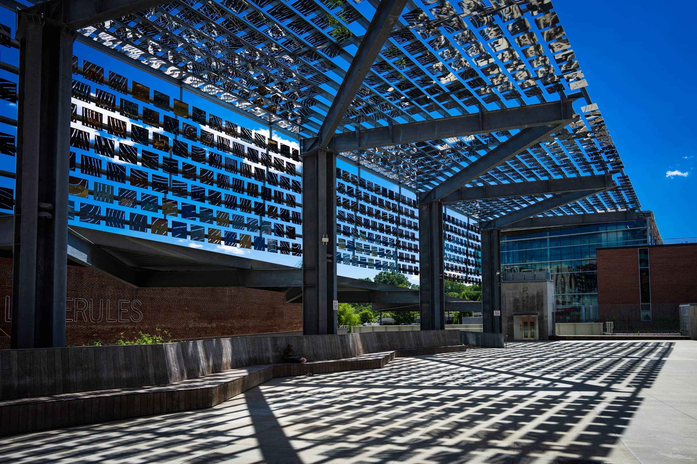 A modern architectural structure reminiscent of Union Station Raleigh, with a grid-like roof casting intricate shadows. The building's brick walls and glass windows create a sleek design, perfect for wedding photography against the clear blue sky.