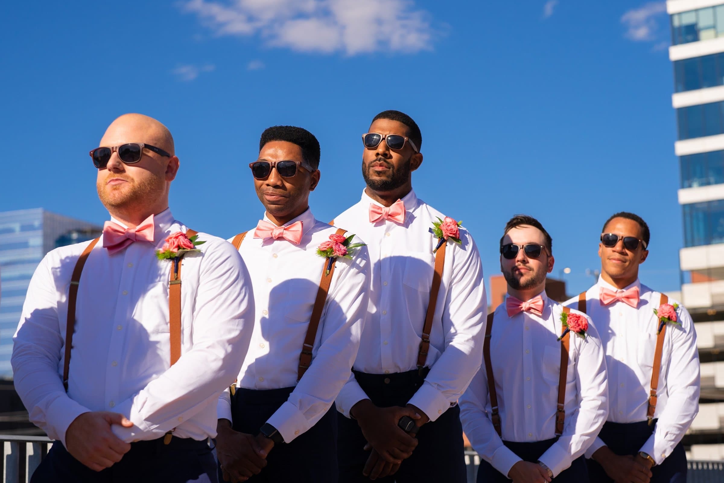 Five groomsmen wearing white shirts, pink bow ties, and suspenders stand in a line outdoors on a sunny day at Union Station Raleigh. They each wear sunglasses and have pink flower boutonnieres. A clear blue sky and modern buildings are in the background, capturing the perfect wedding photography moment.