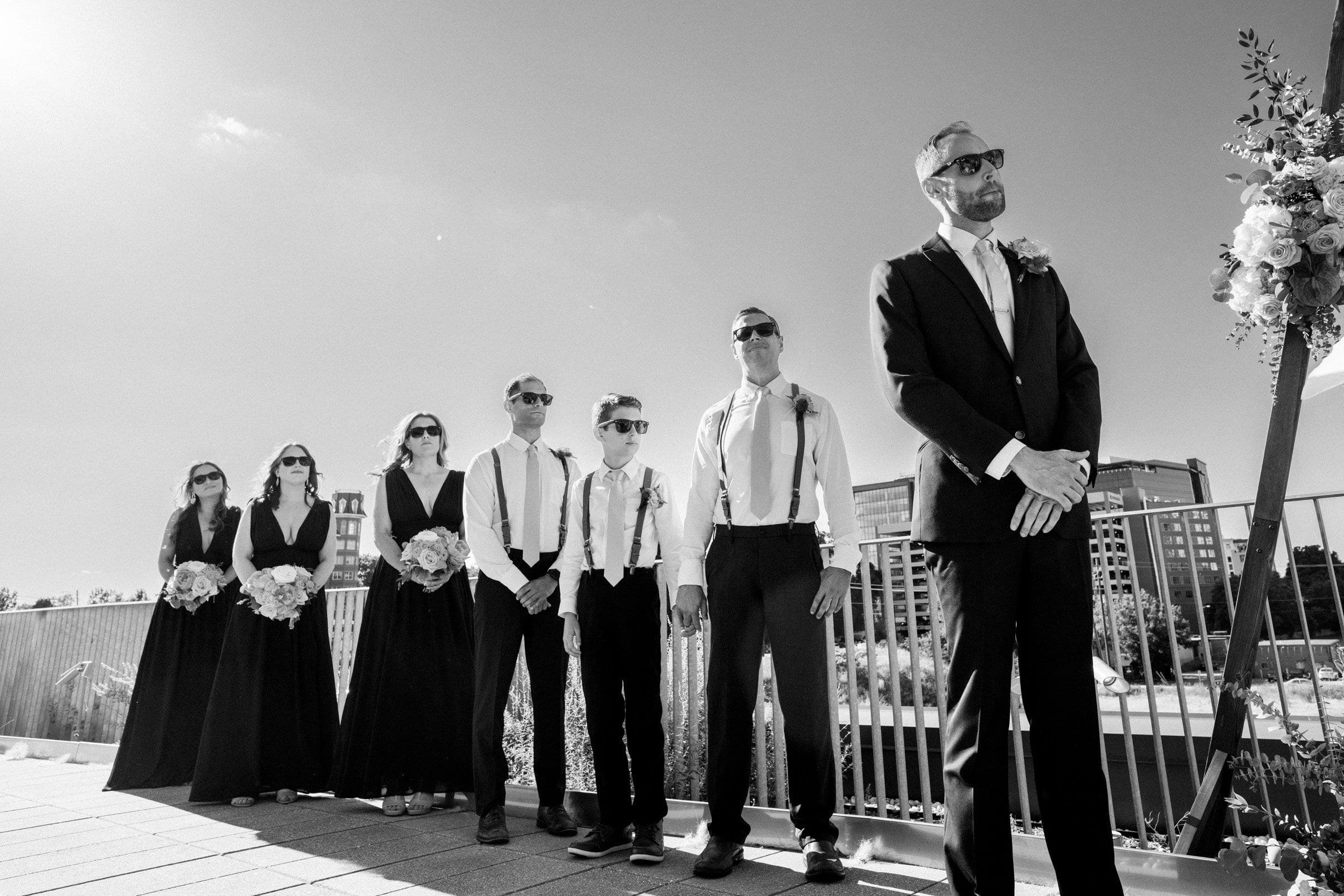 A black-and-white photo captures a Union Station Raleigh wedding party outdoors. Four bridesmaids in dark dresses and three groomsmen in white shirts with suspenders and sunglasses stand beside the groom in a dark suit, with a cityscape visible under the clear sky.