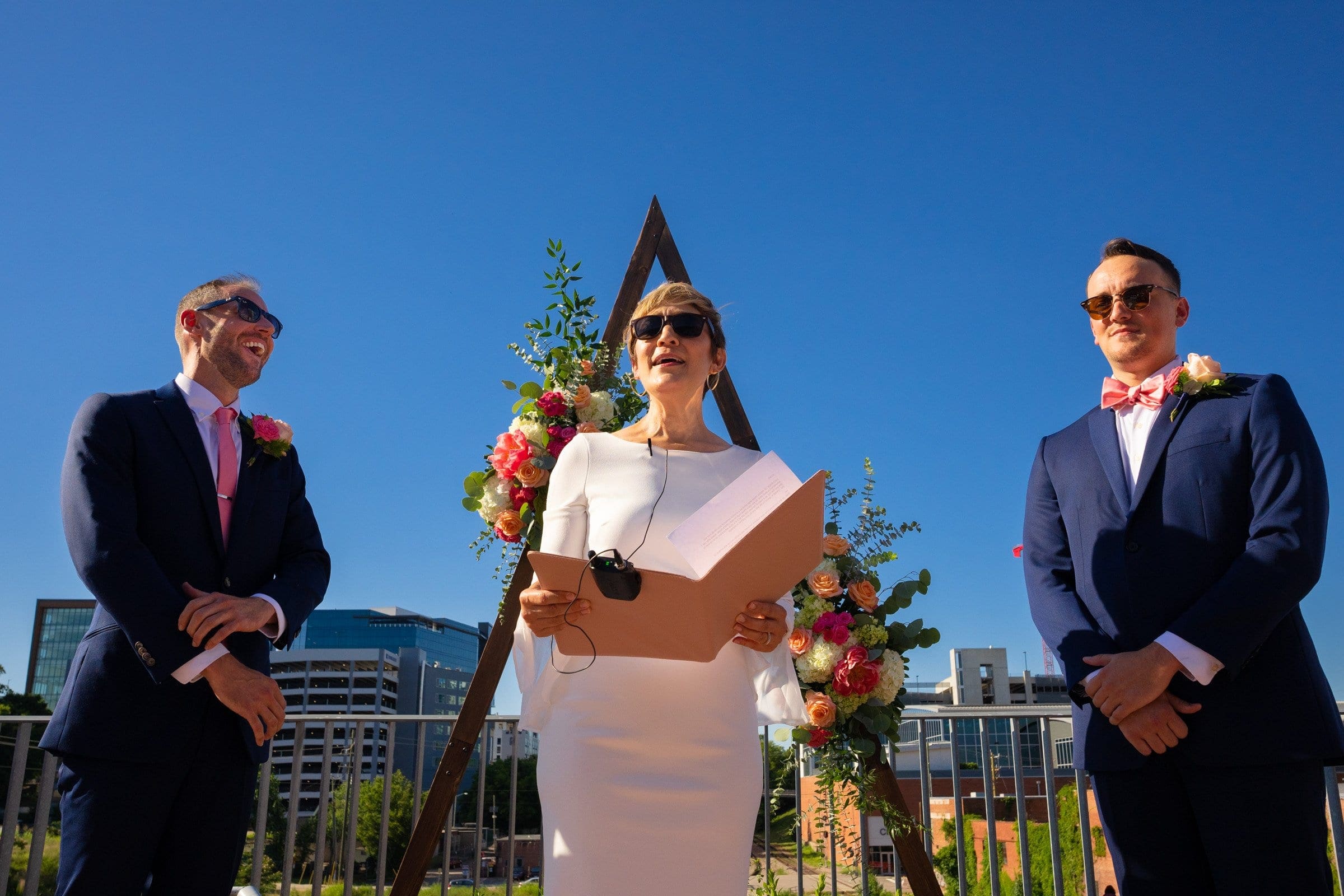 Three people at an outdoor wedding. One person in a white dress holds a paper and speaks, standing between two people in suits with pink boutonnieres. With the bright blue sky and cityscape reminiscent of Union Station Raleigh, it’s a perfect moment captured through wedding photography.